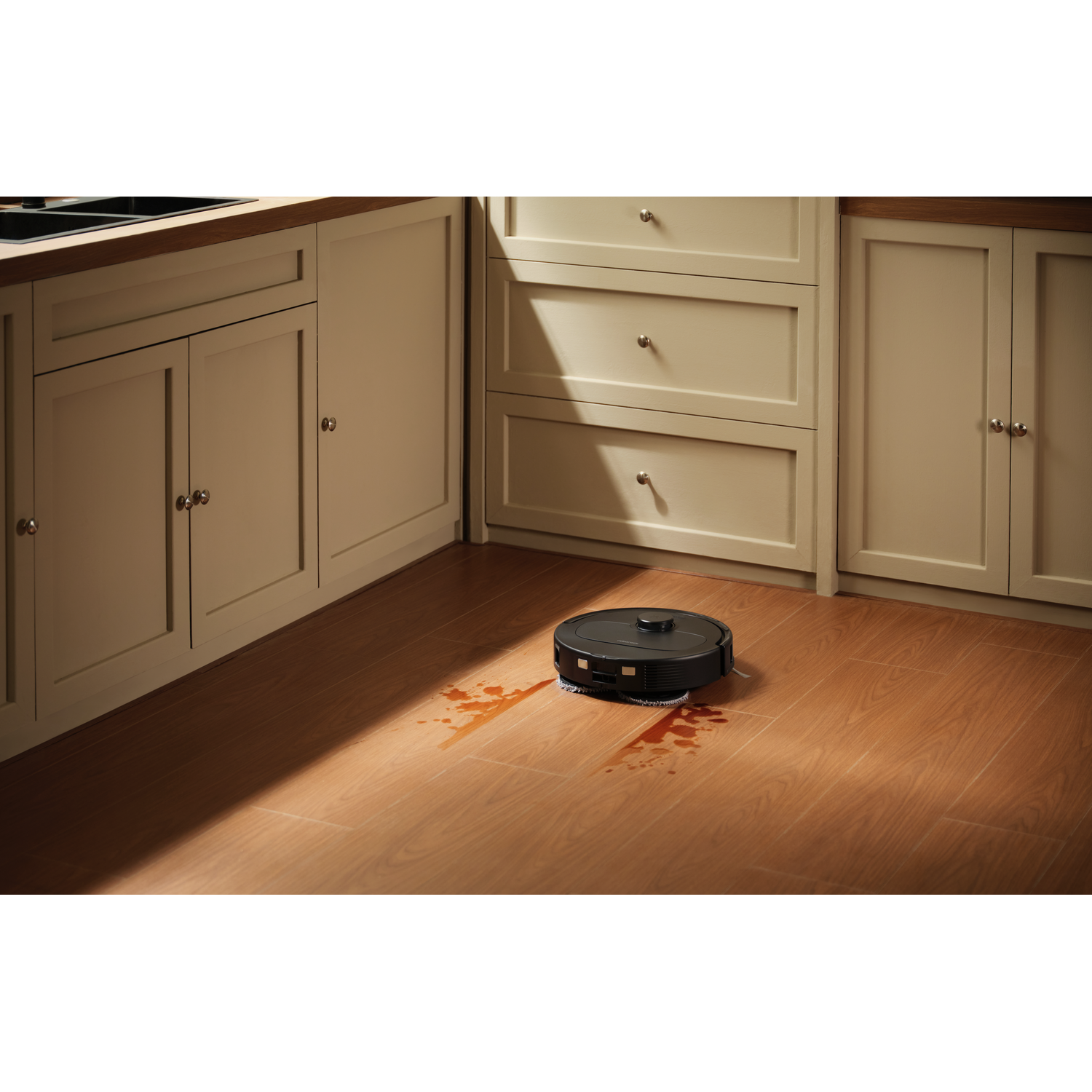 Un robot aspirateur noir nettoie un plancher en bois dans une cuisine lumineuse avec des taches de café.
