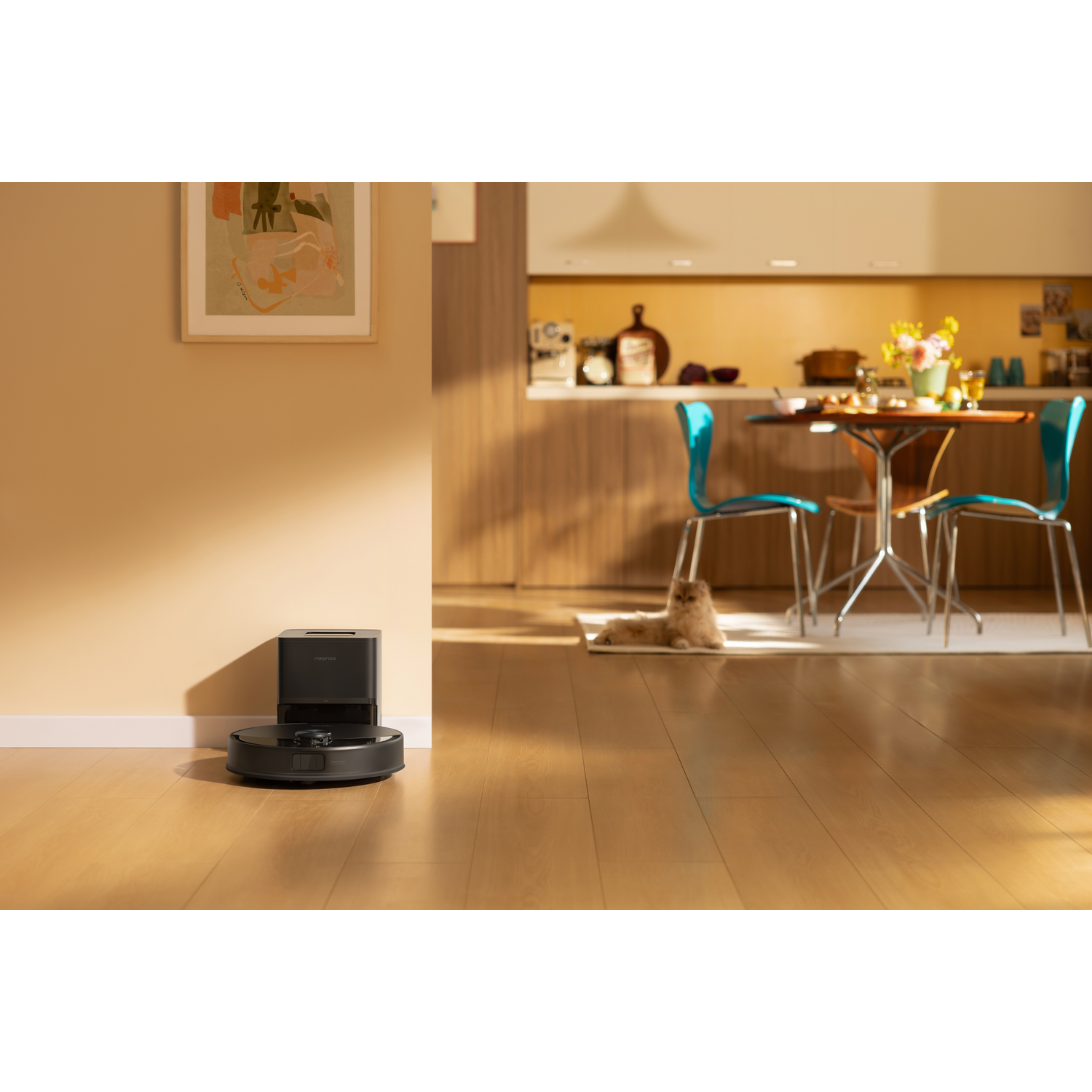 Un aspirateur robot noir se recharge sur son socle en bois, avec une cuisine et un chat en arrière-plan.