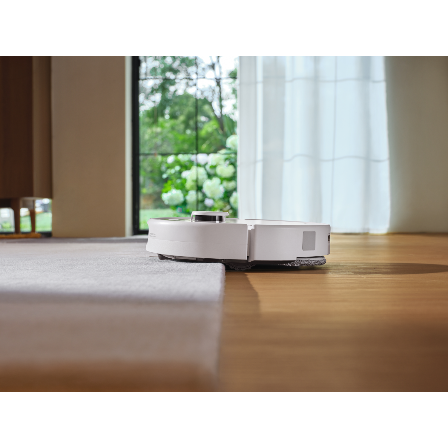 Un robot aspirateur blanc passe du parquet à un tapis clair dans une pièce ensoleillée.