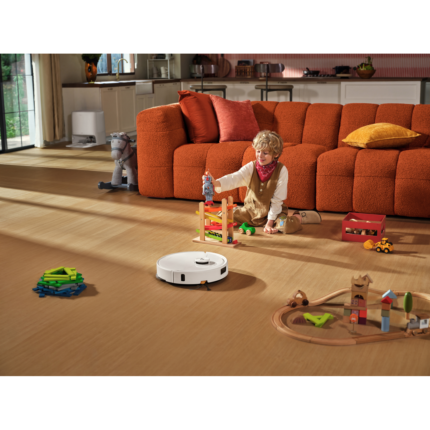 Un enfant joue avec des jouets au sol pendant qu'un robot aspirateur nettoie dans un salon ensoleillé.