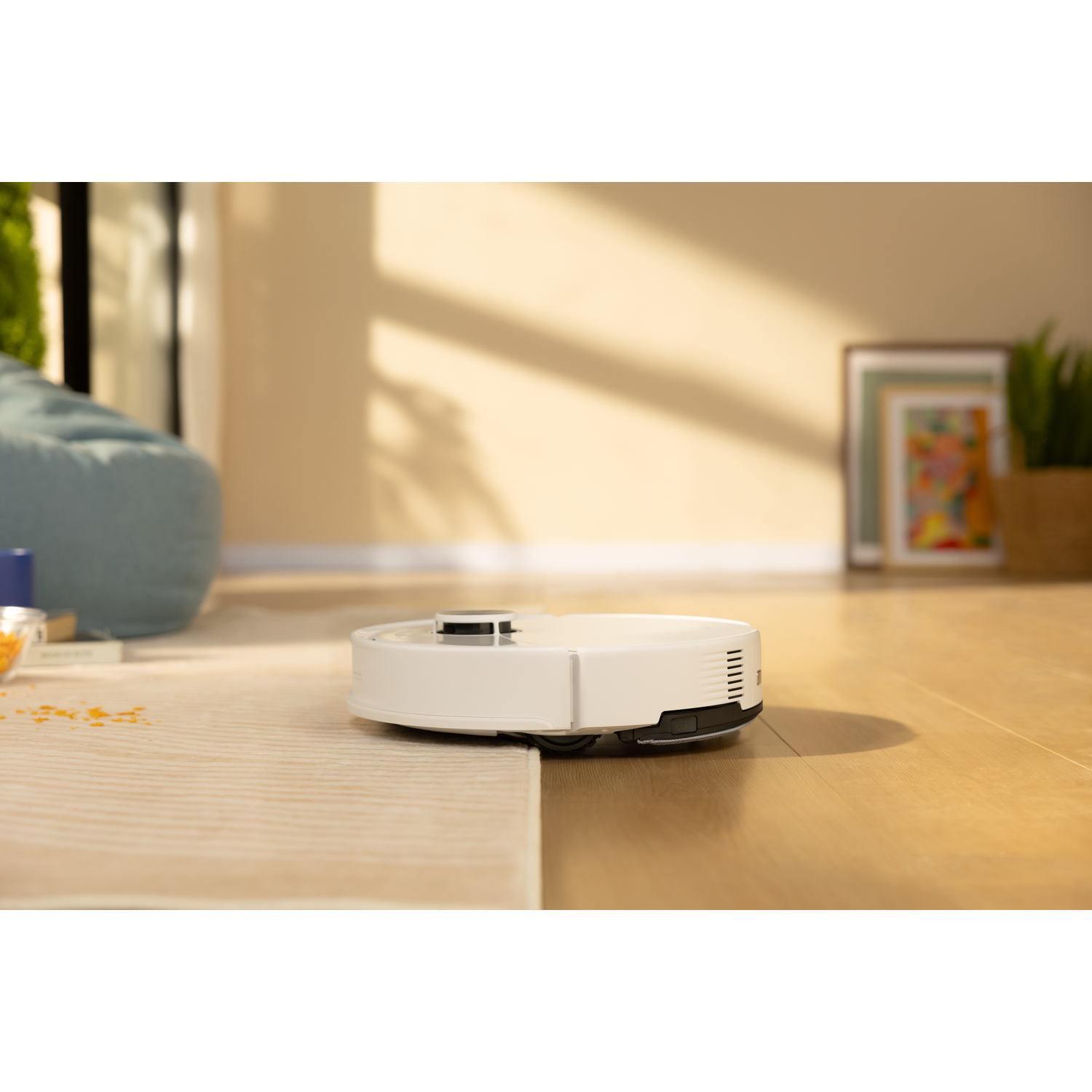 Un robot aspirateur blanc passe d'un sol en bois à un tapis clair dans une pièce ensoleillée.