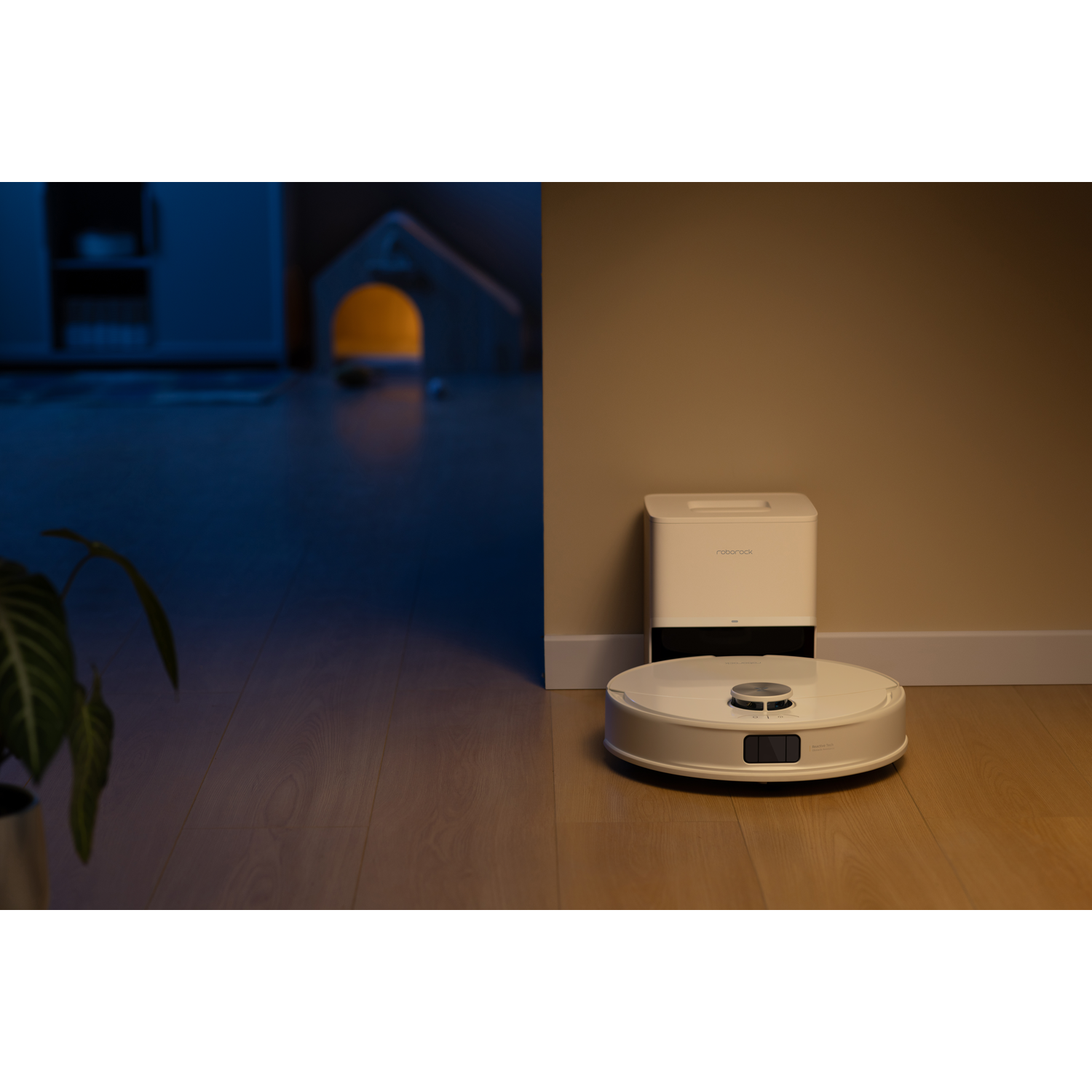 Un robot aspirateur blanc s'arrime à sa station de charge sur un sol en bois dans une pièce tamisée.