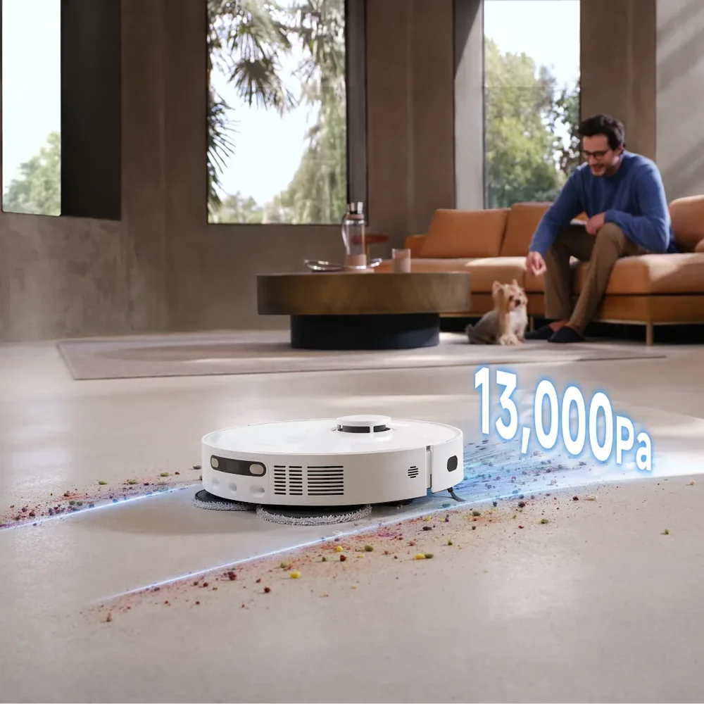 Egy fehér robotporszívó takarítja a padlót, miközben a háttérben egy férfi és egy kutya ül a kanapén.