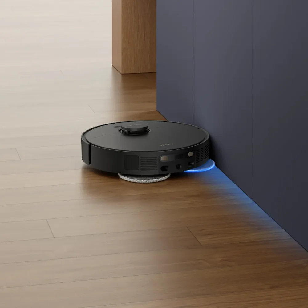 Egy fekete robotporszívó tisztít egy fa padlót kék szekrények mellett, kékes fényt bocsátva ki.