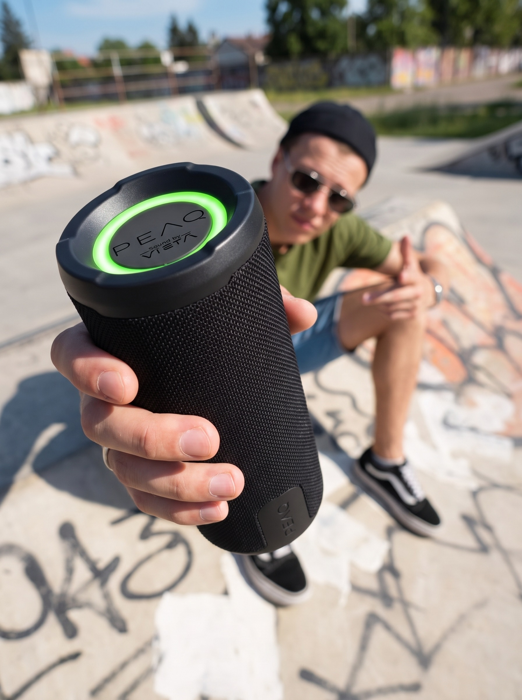 Un hombre en un skatepark sostiene un altavoz Bluetooth negro cilíndrico con un anillo de luz verde hacia la cámara.