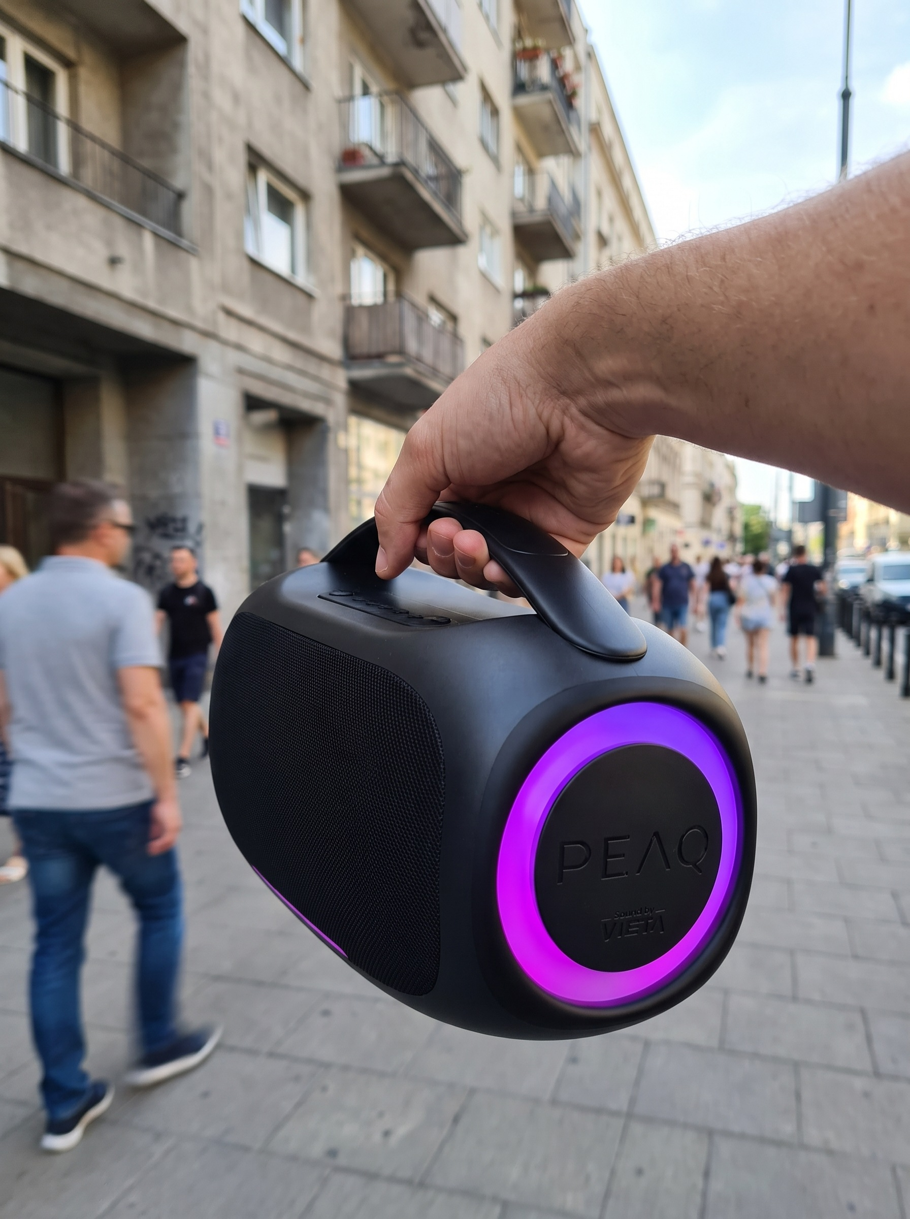 Mano sosteniendo un altavoz Bluetooth PEAQ negro con aro de luz violeta.