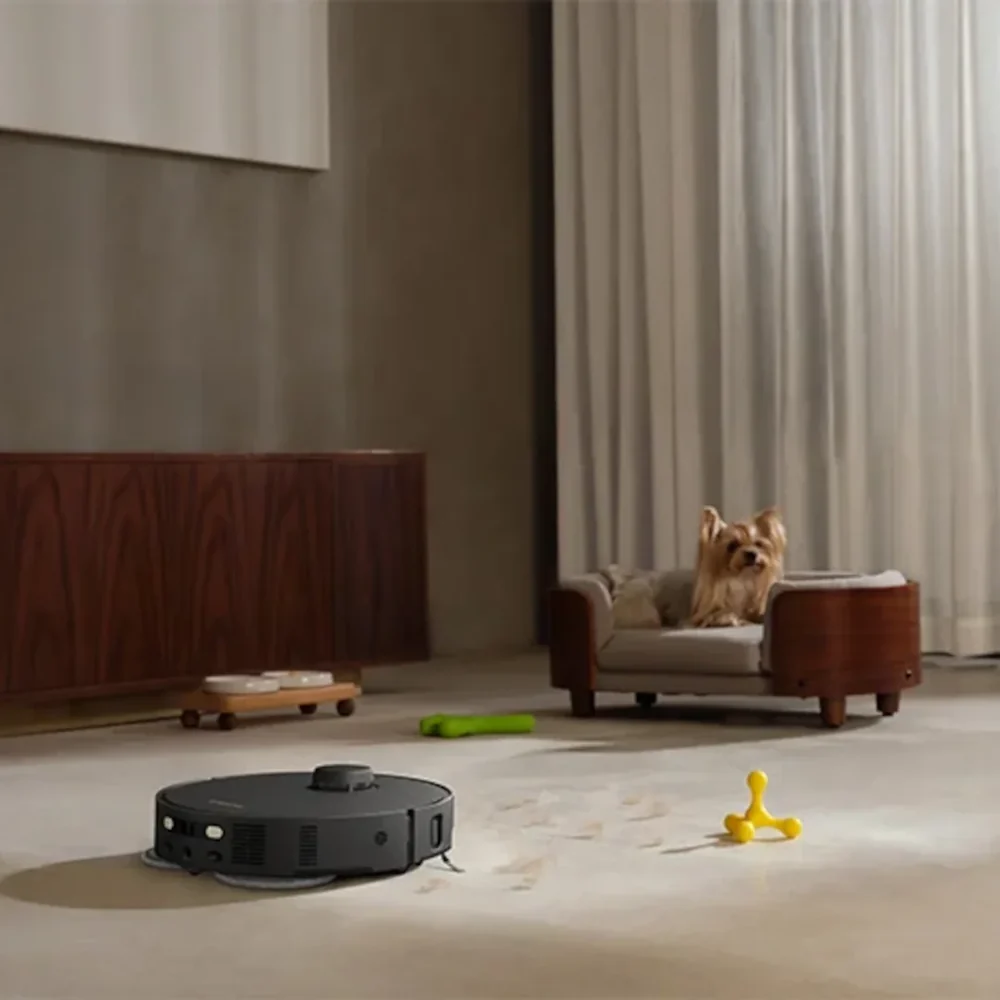 Egy robotporszívó takarítja a padlót, míg egy kis kutya a fekhelyén ül.