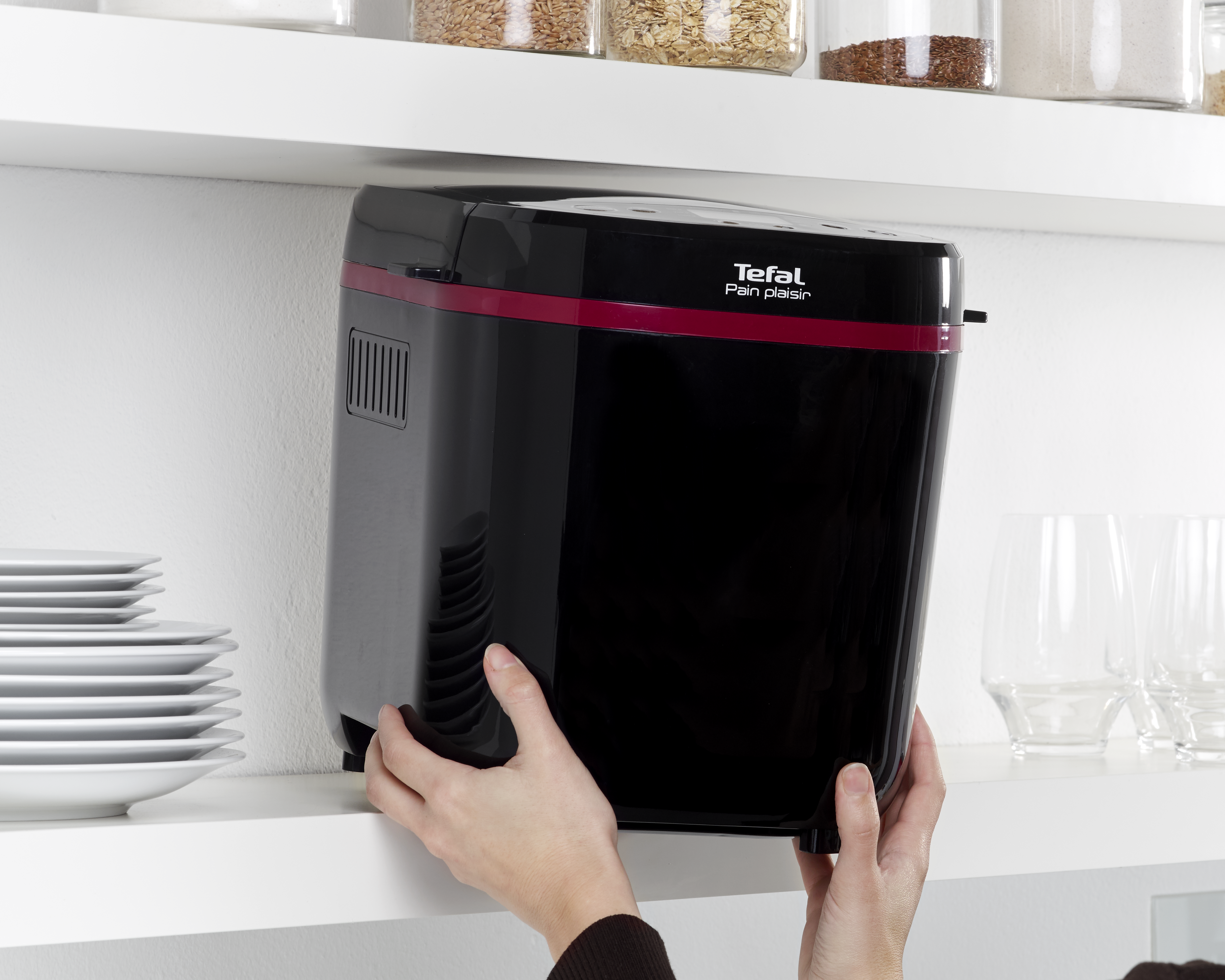 Kezek egy fekete Tefal kenyérsütőt helyeznek el egy konyhai polcon tányérokkal és poharakkal.