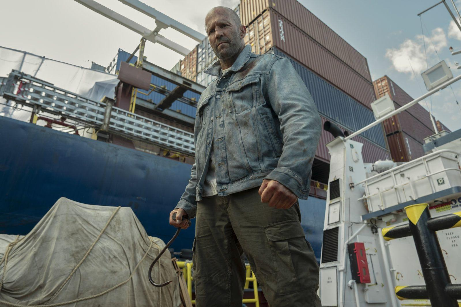 Ein Mann im grauen Jeanshemd und Cargo-Hose hält einen Haken auf einem Schiff, im Hintergrund sind Frachtcontainer.