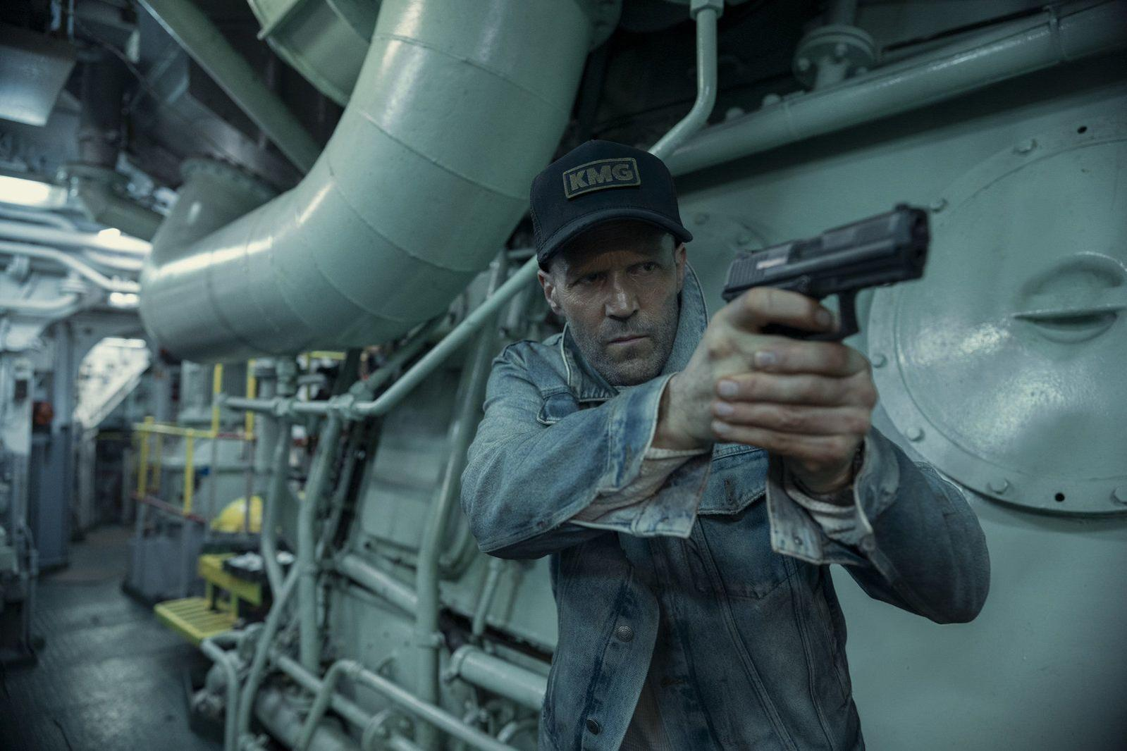 Ein Mann im Denim-Hemd und Kappe zielt mit einer Pistole in einem industriellen Raum mit großen Rohren.