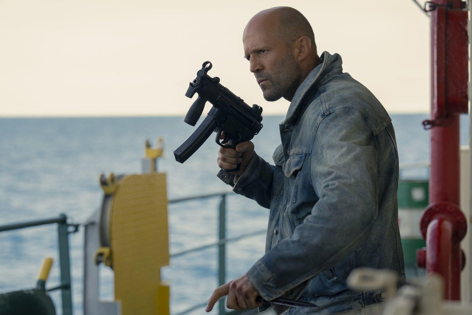 Ein kahlköpfiger Mann in einer Jeansjacke hält auf einem Schiff eine Maschinenpistole.