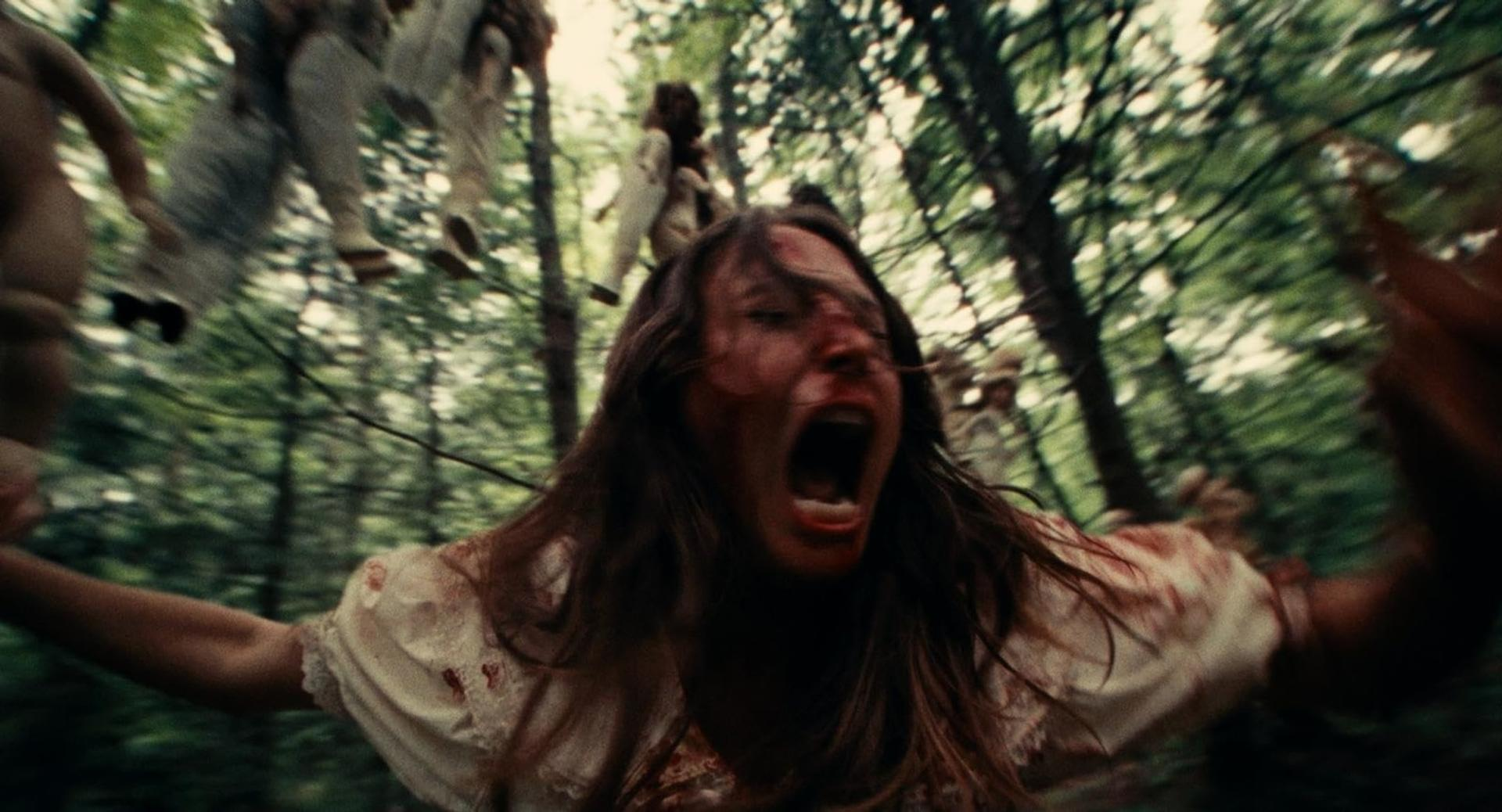 Eine blutige Frau schreit panisch in einem Wald, im Hintergrund hängen Puppen in den Bäumen.