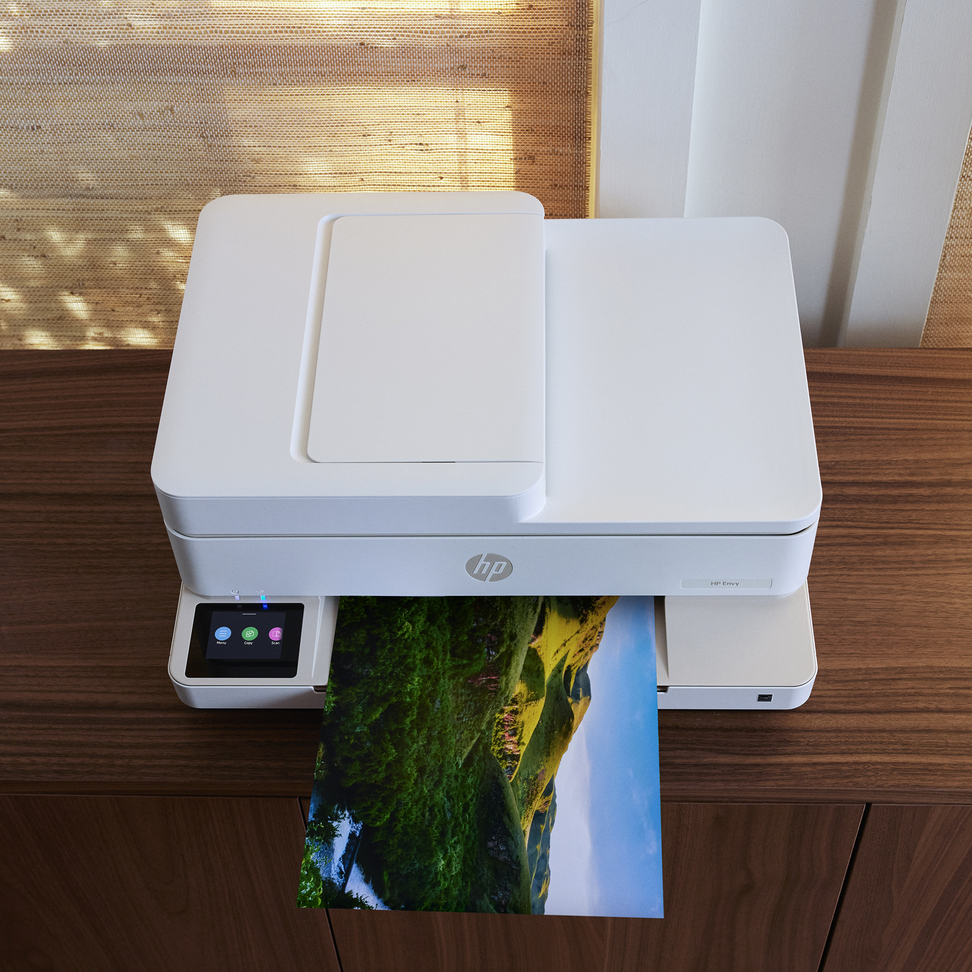 Ein weißer HP Envy Drucker steht auf einem Holzschrank und druckt ein Bild einer grünen Landschaft.
