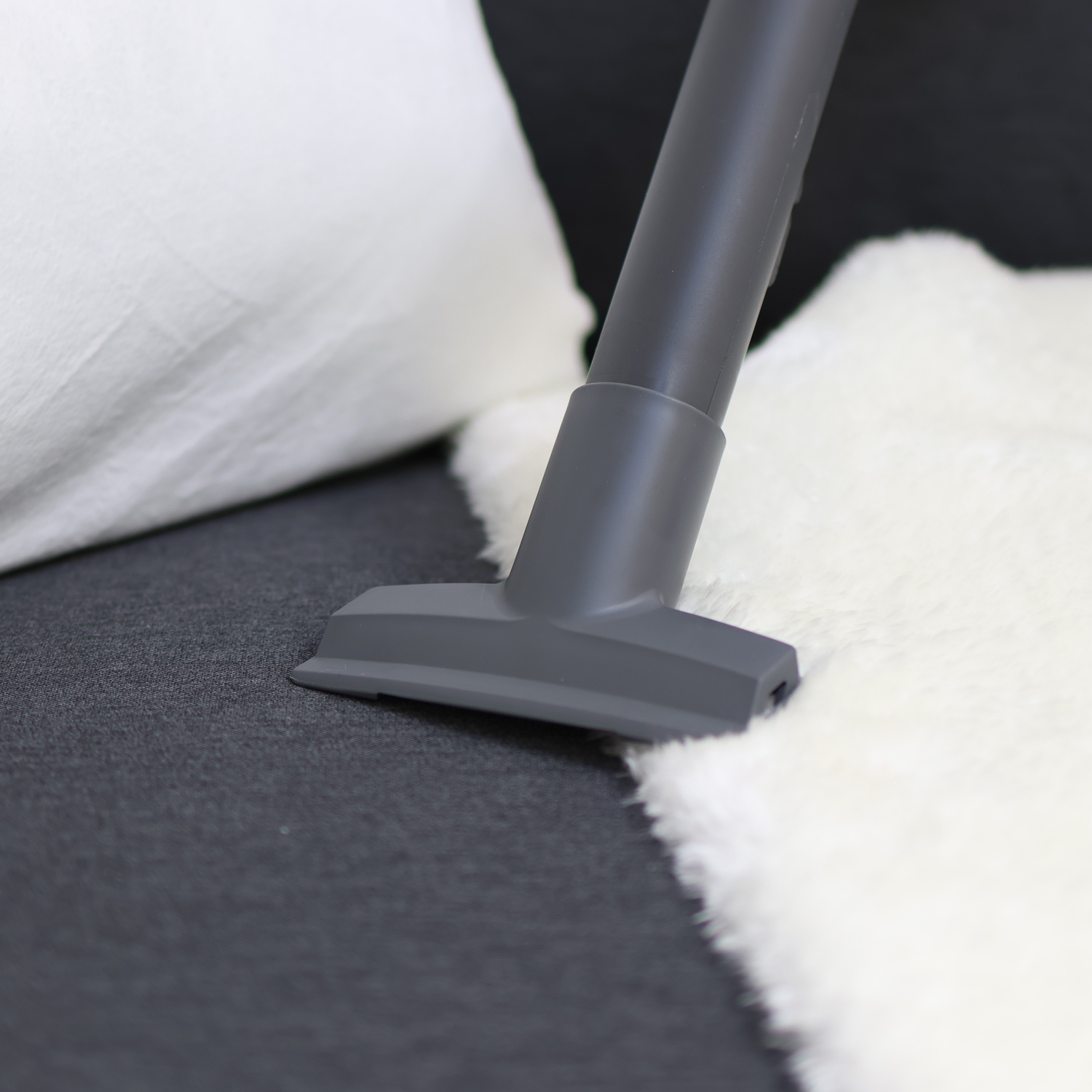 Ein grauer Staubsaugeraufsatz reinigt eine weiße flauschige Decke auf einem dunklen Sofa.