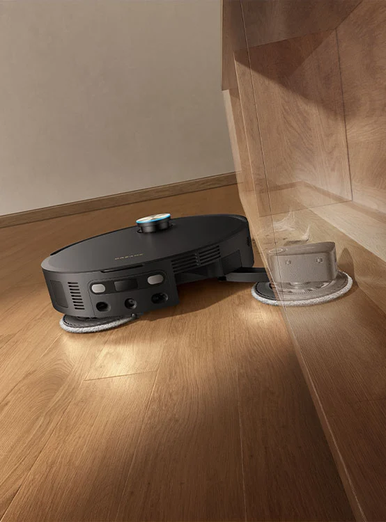 Egy fekete robotporszívó tisztítja a fa padlót egy bútor alatt.