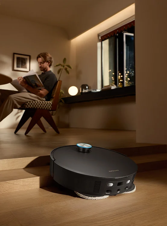 Egy fekete robotporszívó tisztítja a fapadlót, míg egy férfi könyvet olvas a háttérben lévő nappaliban.