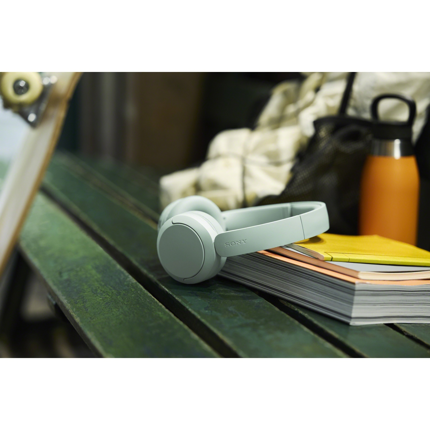 Casque Sony vert clair posé sur des cahiers sur un banc en bois, avec une bouteille orange à côté.