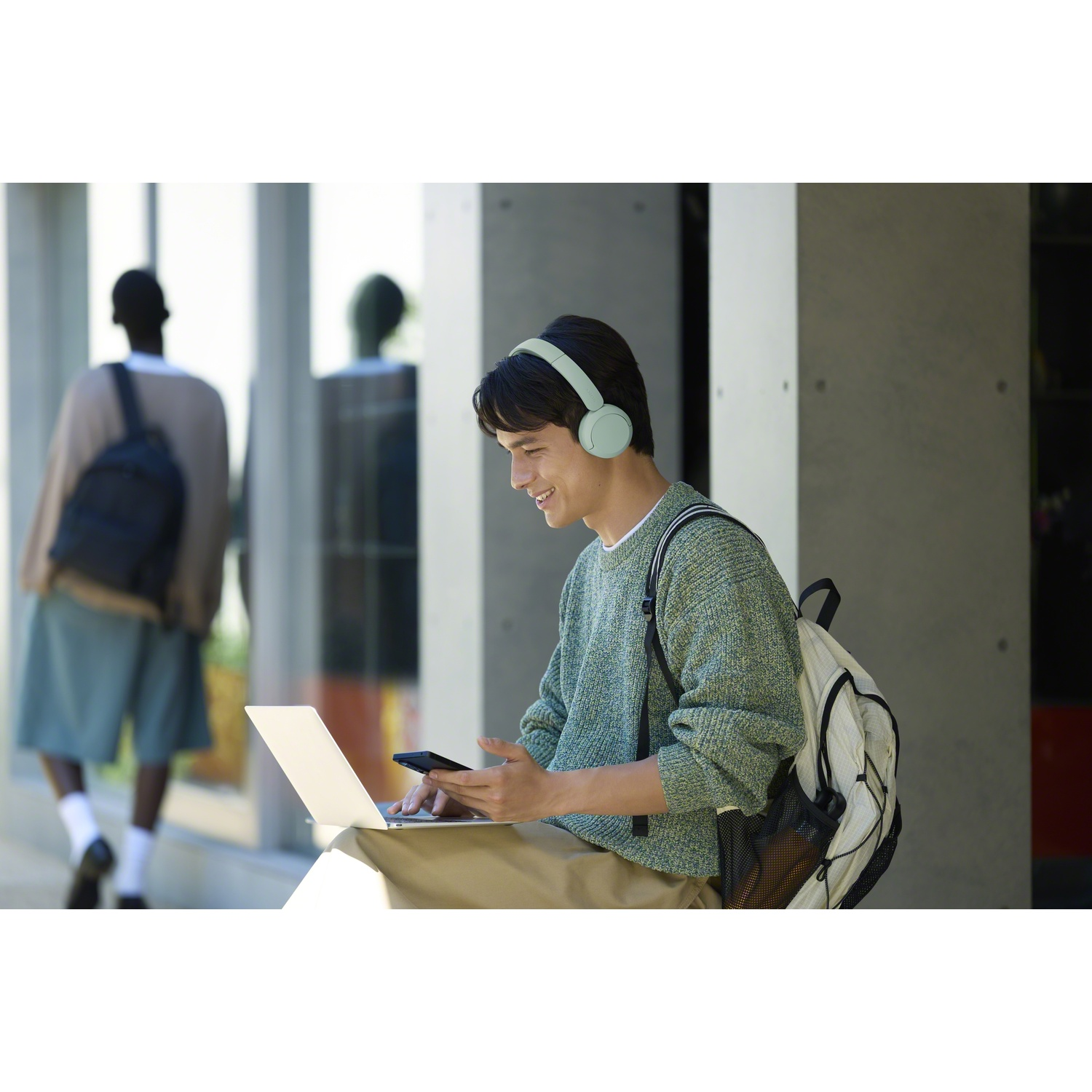 Un jeune homme avec un casque et un sac à dos est assis dehors, utilisant un ordinateur et un smartphone.
