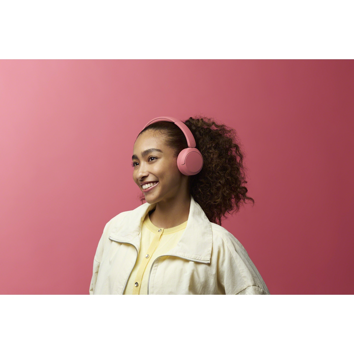Jeune femme souriante avec un casque rose et une veste beige sur fond rose.