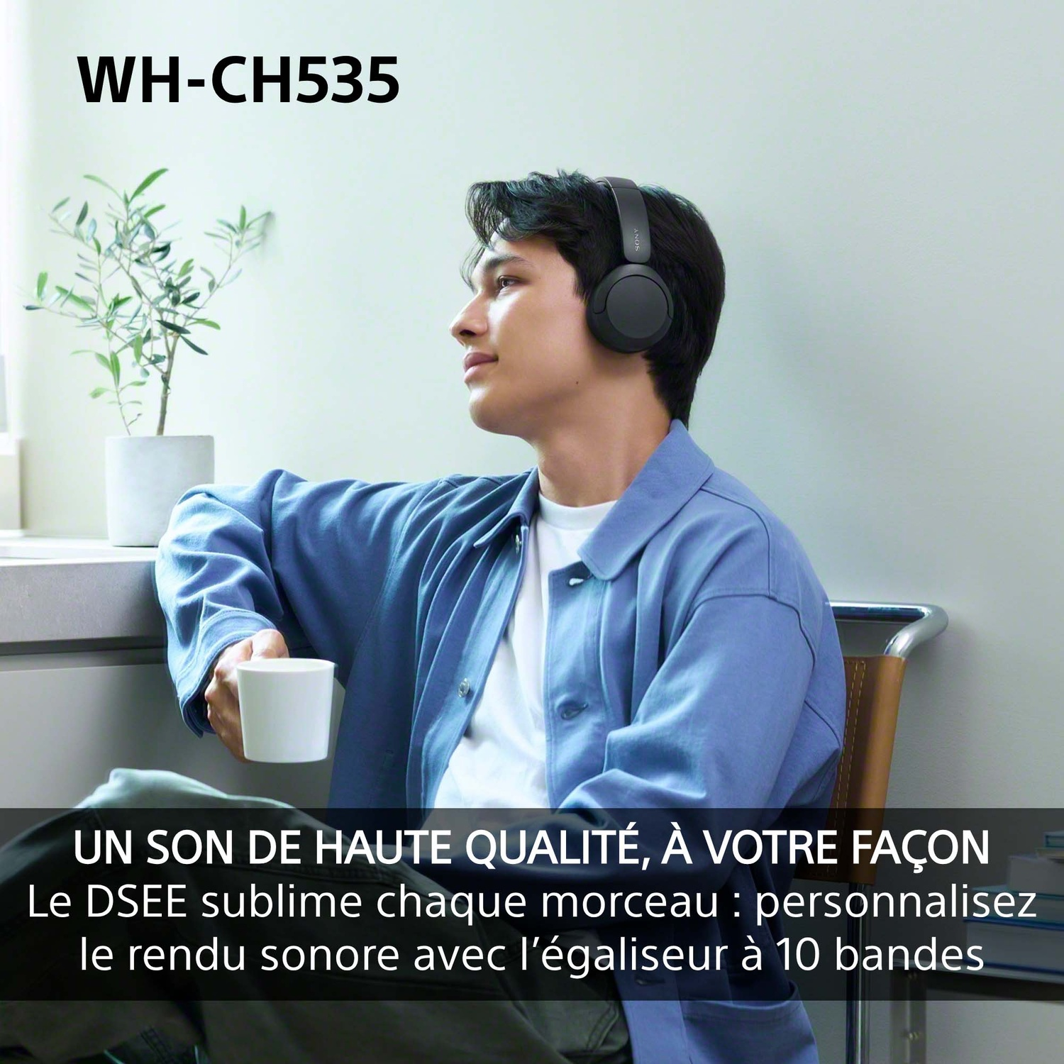 Un jeune homme portant un casque noir tenant une tasse blanche, assis à l'intérieur avec une plante en arrière-plan.