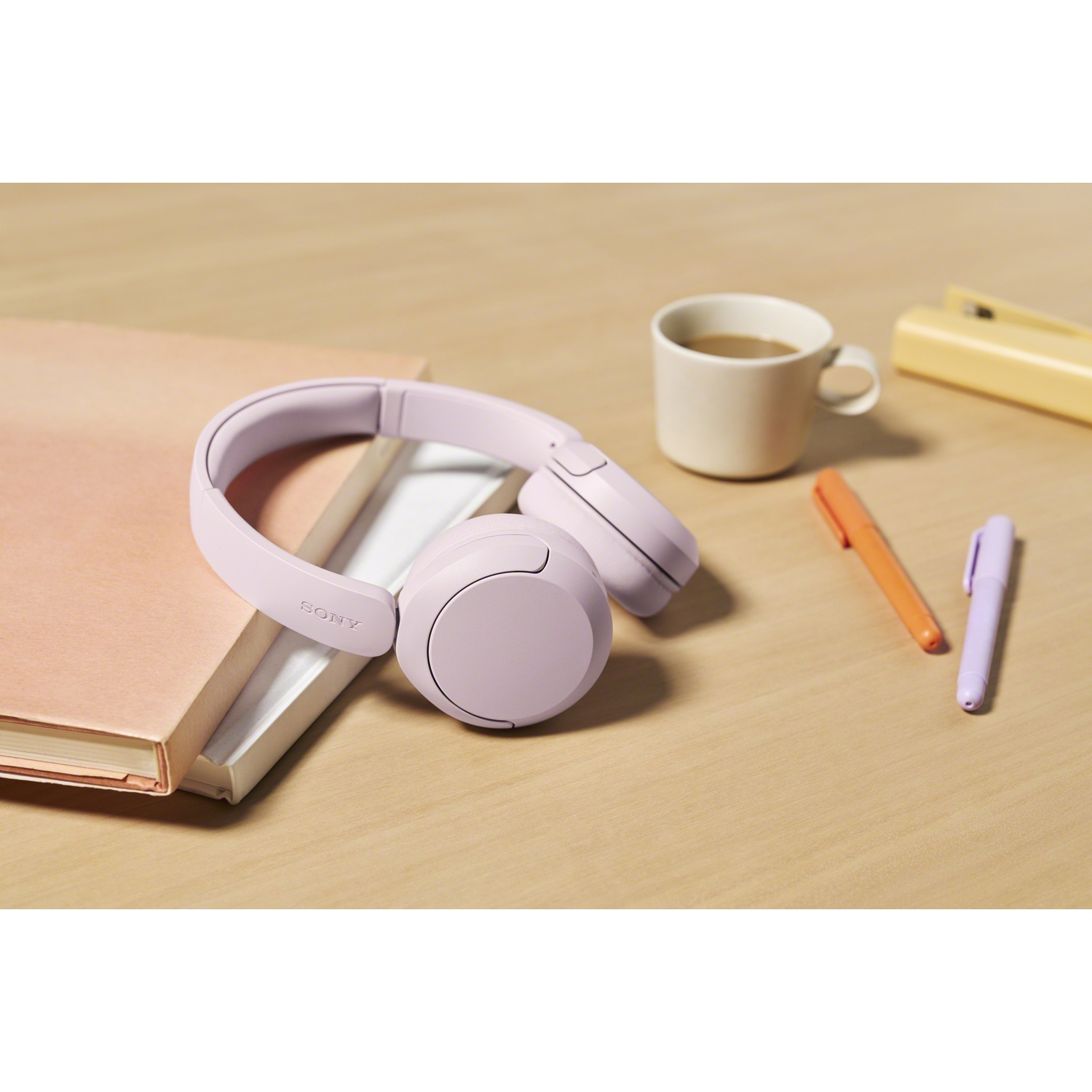 Casque lilas Sony sur des livres près d'une tasse de café et de stylos sur un bureau en bois.