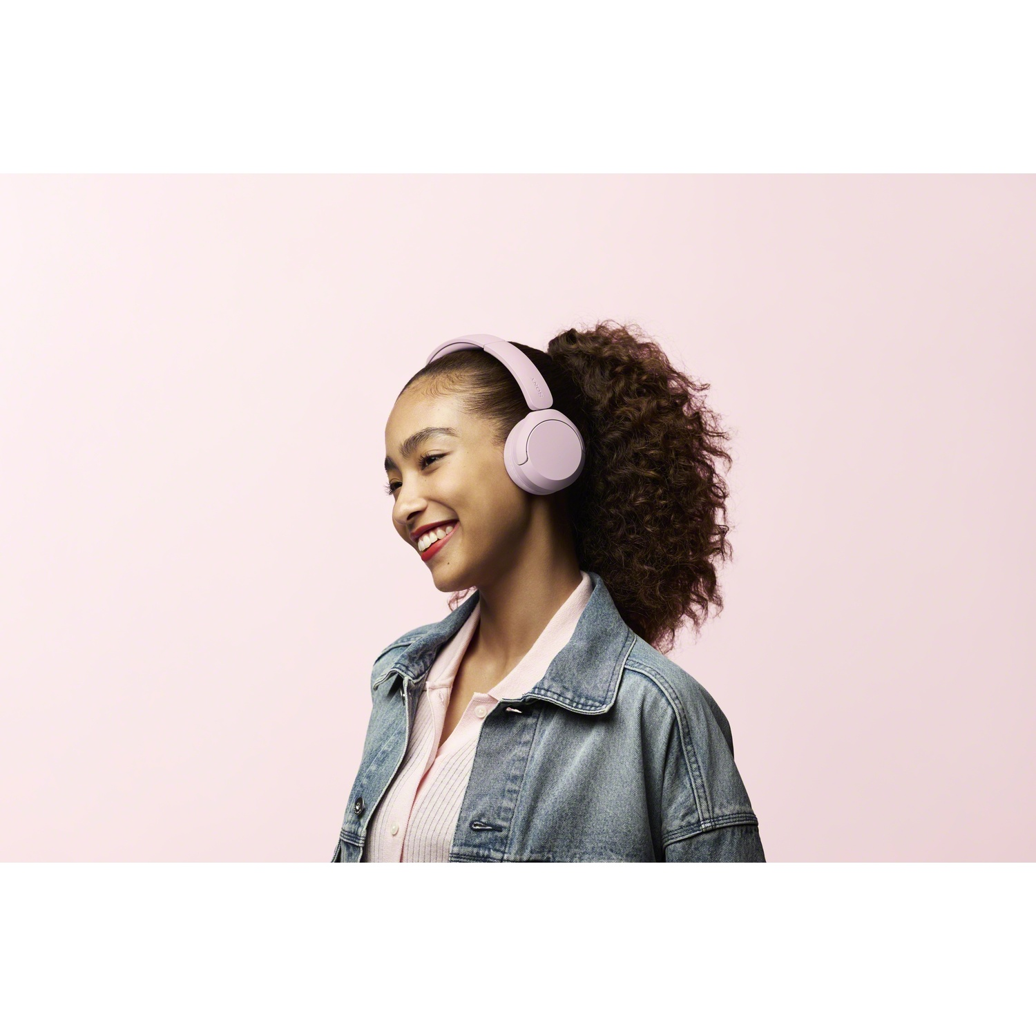 Une femme souriante aux cheveux bouclés porte un casque violet et une veste en jean sur fond rose.