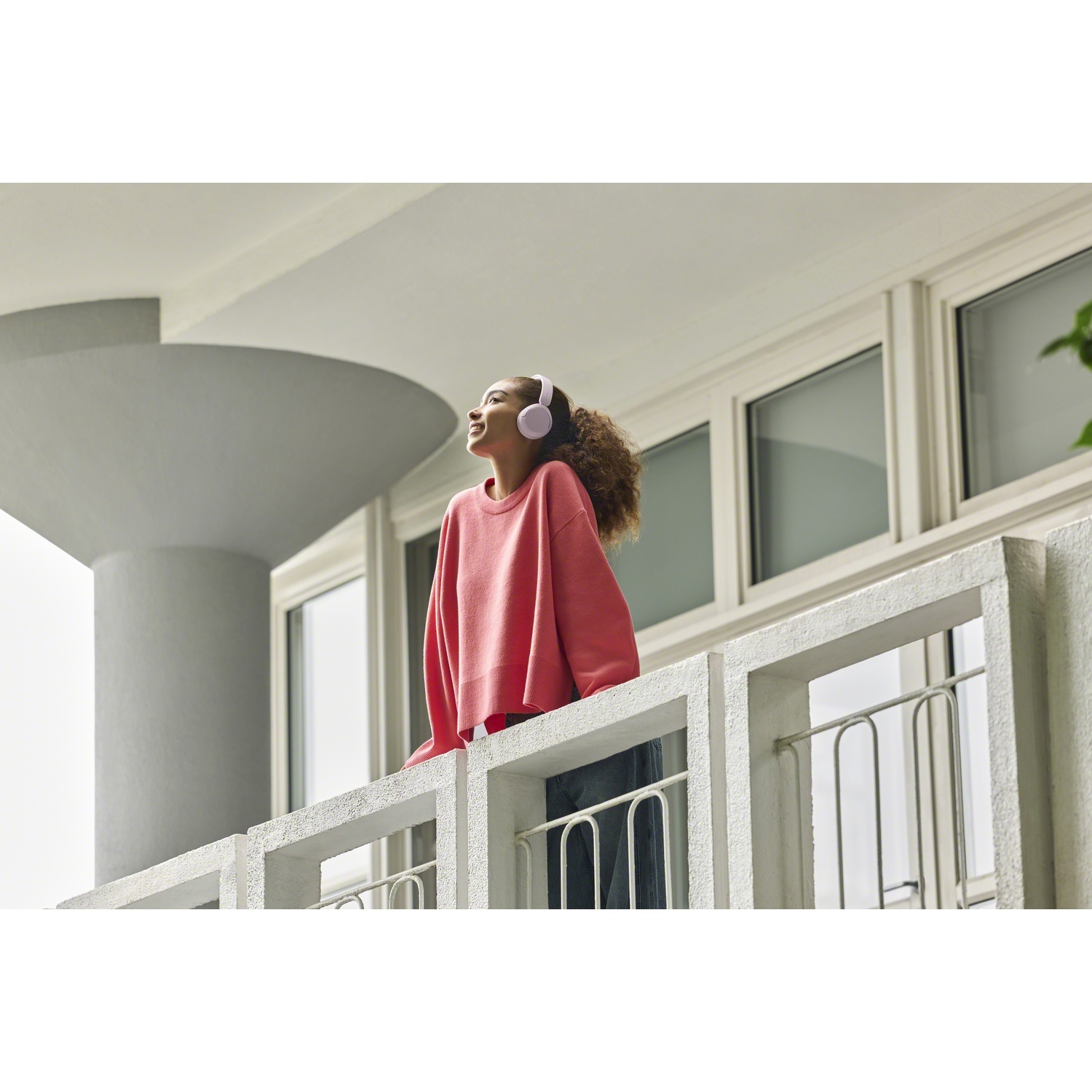 Une jeune femme en pull rose portant un casque se tient sur un balcon et lève les yeux.