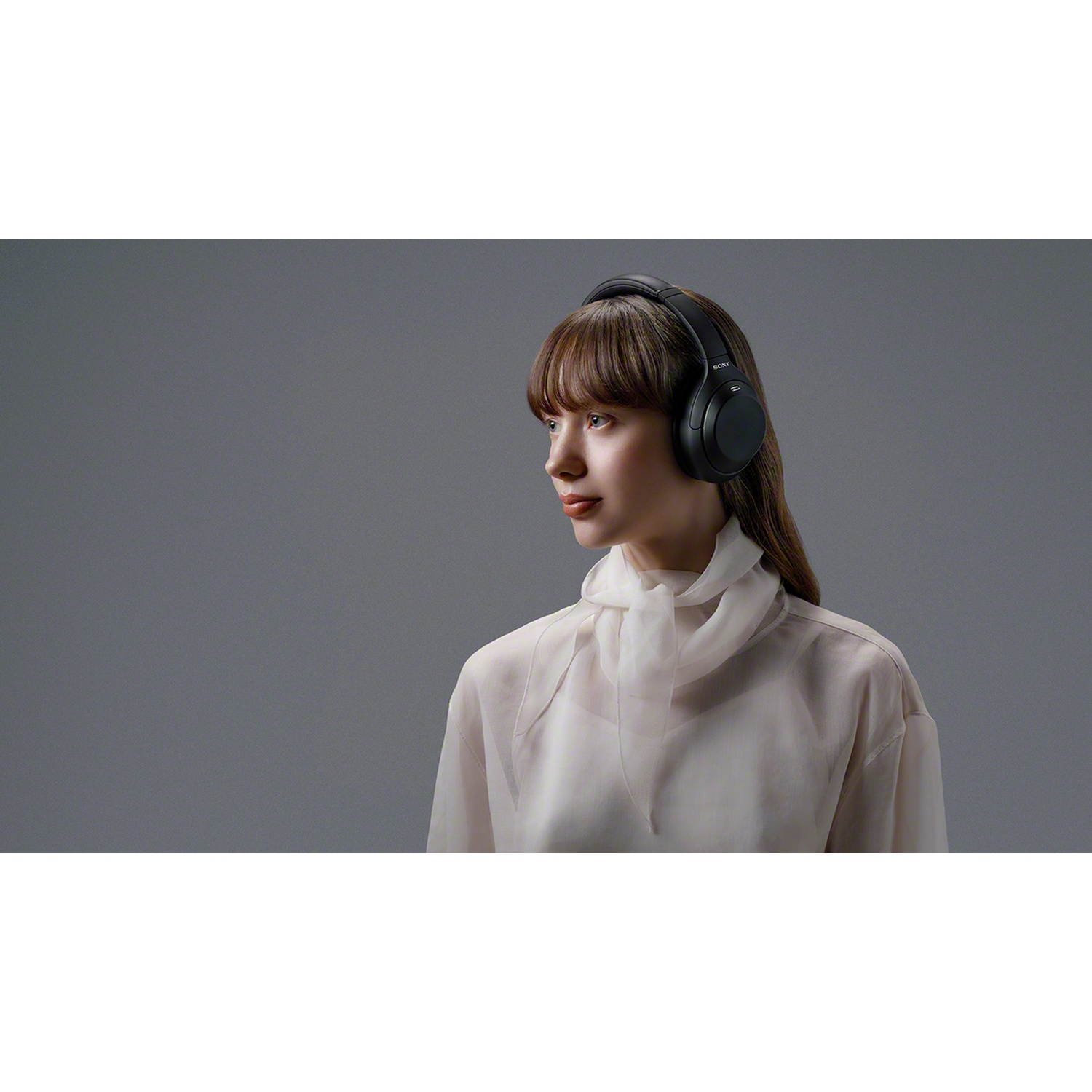 Une femme aux cheveux bruns porte un casque noir et un chemisier blanc sur fond gris.