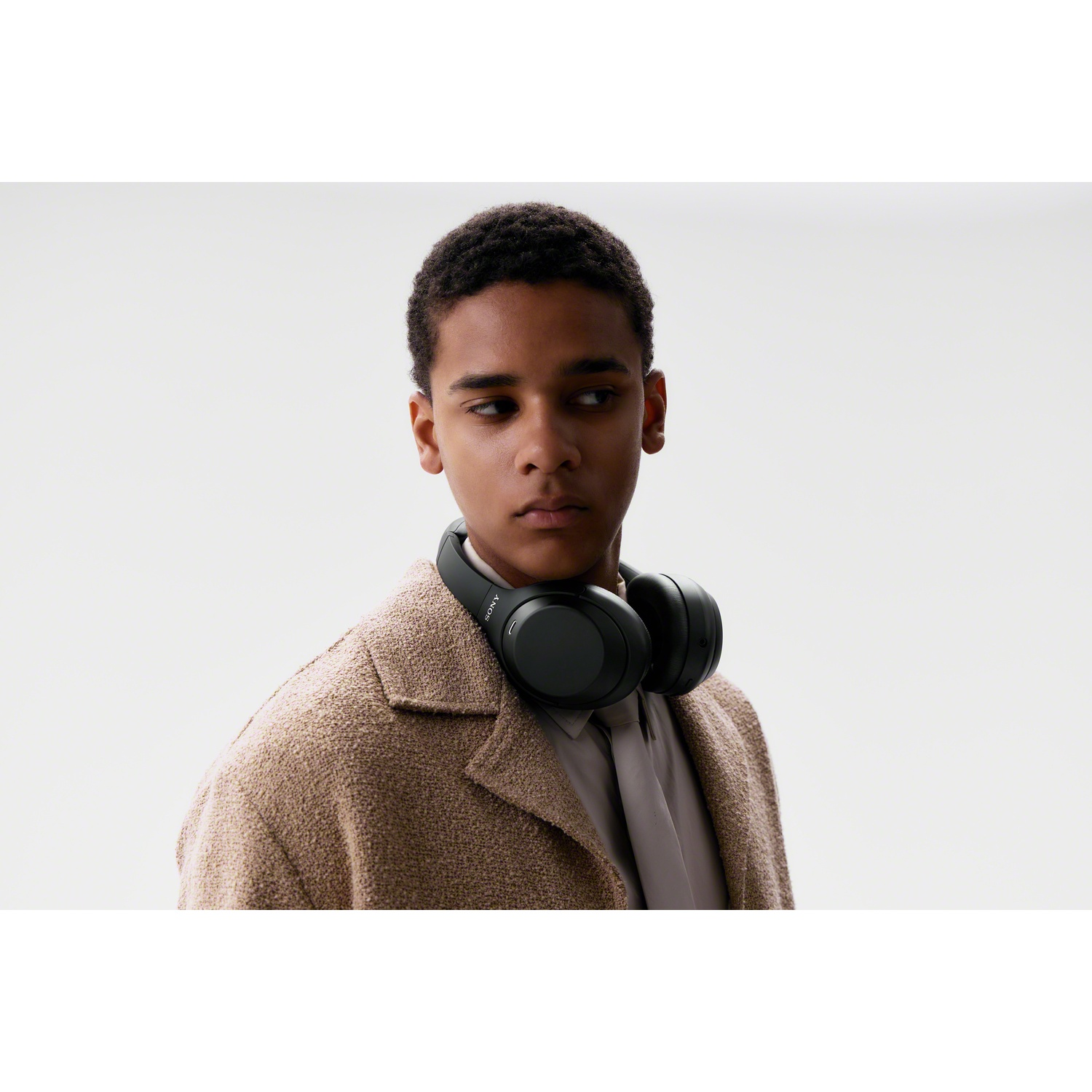Un jeune homme en manteau beige et casque noir autour du cou regarde vers la droite sur fond clair.