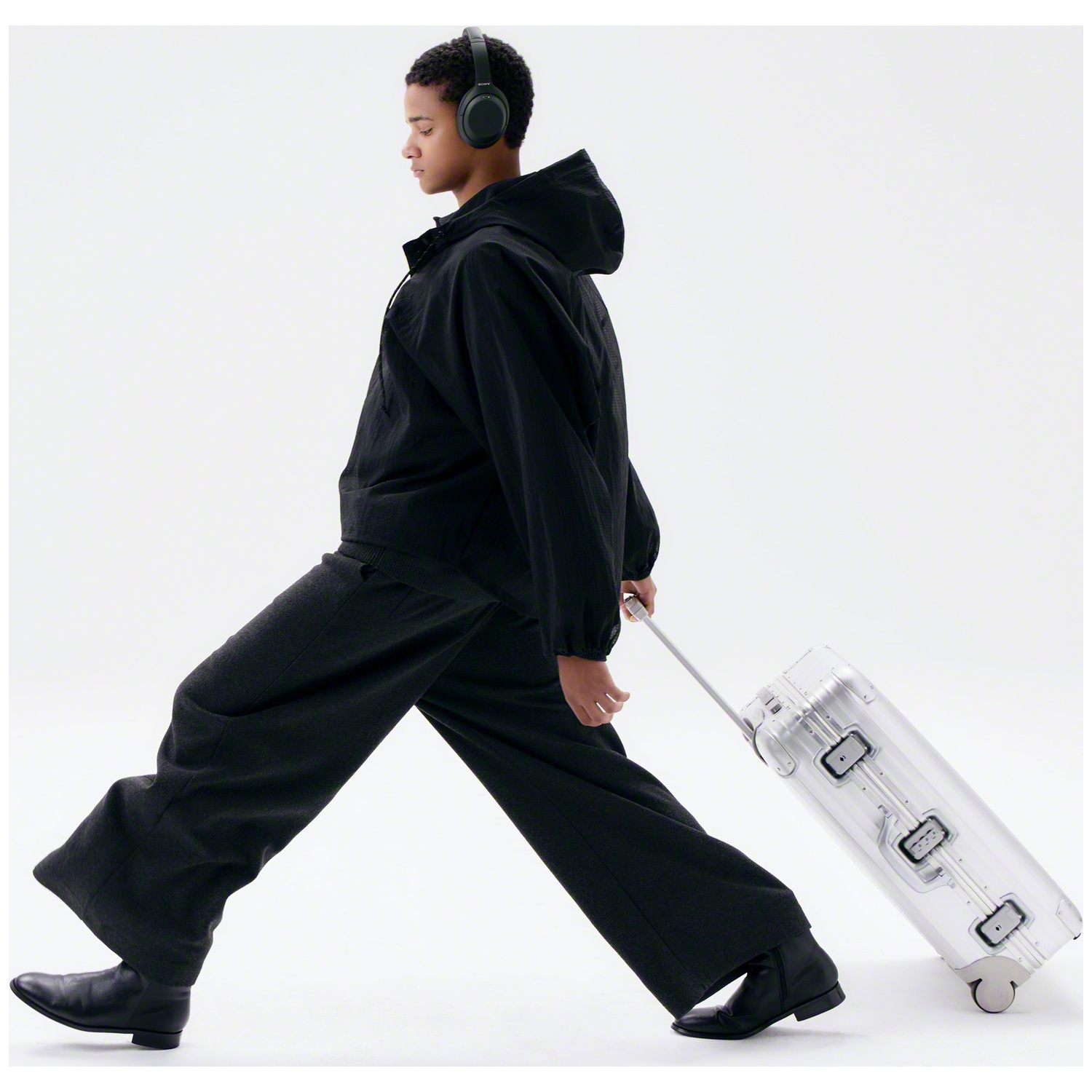 Un jeune homme en noir avec un casque marche et tire une valise argentée sur fond blanc.