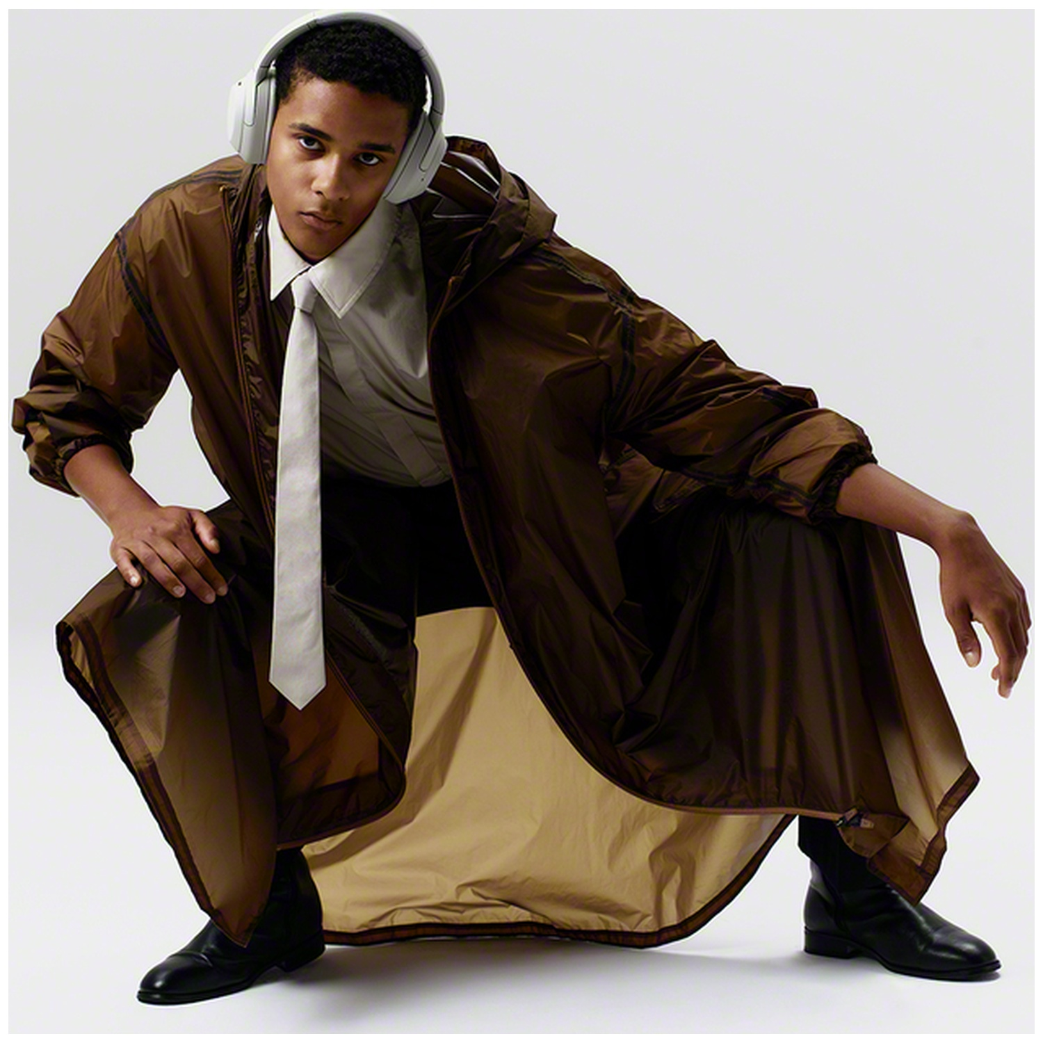 Un jeune homme en manteau marron, chemise blanche, cravate et casque est accroupi sur fond blanc.