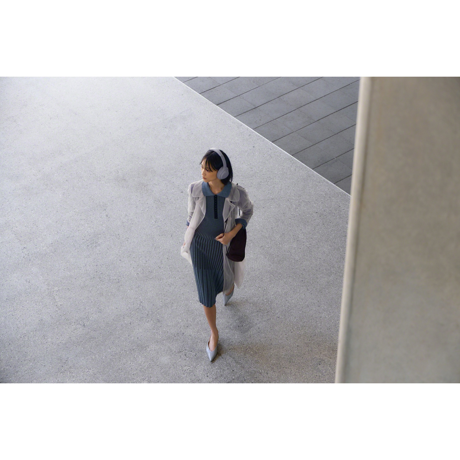 Une femme en robe bleue et manteau marche sur un trottoir gris avec un casque audio.