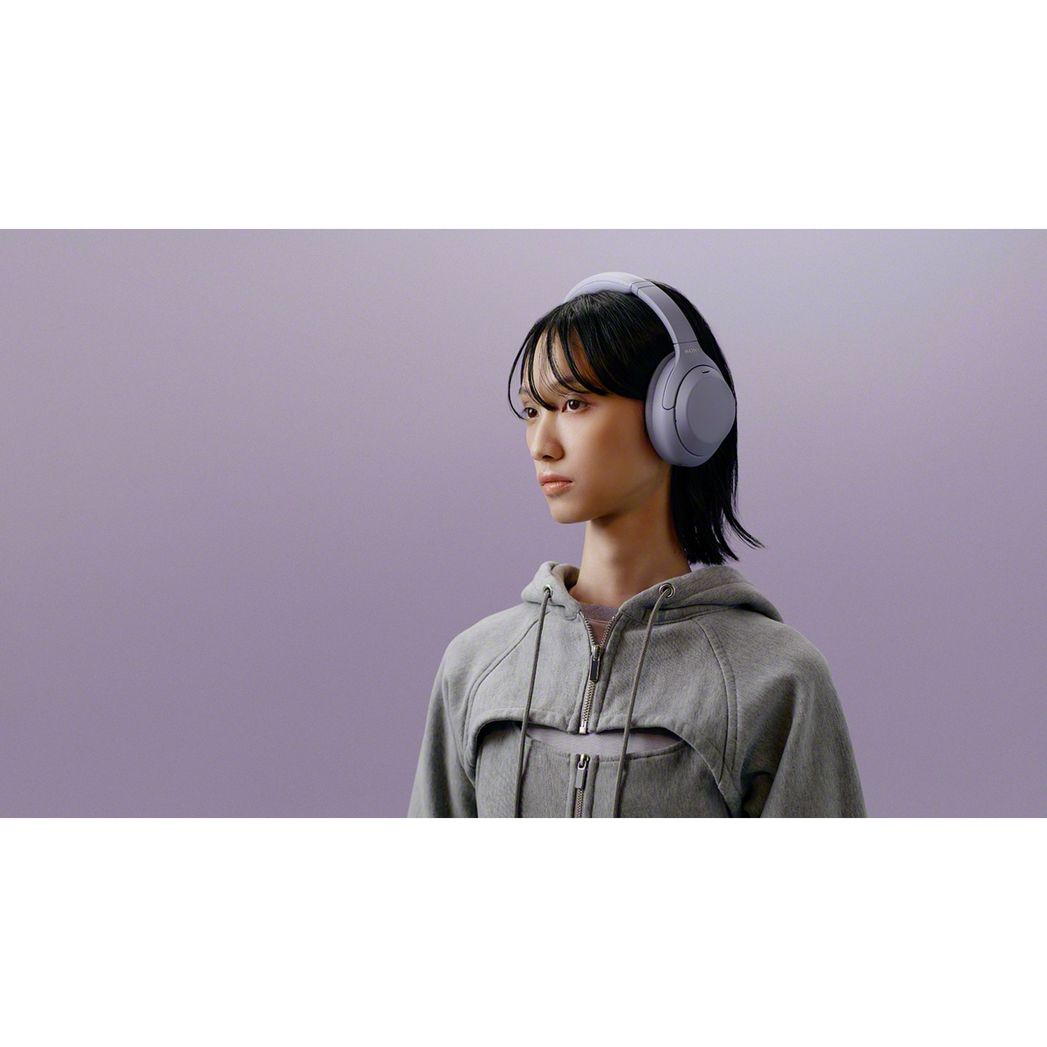 Une jeune femme aux cheveux noirs porte un casque gris et un sweat à capuche gris sur fond mauve.