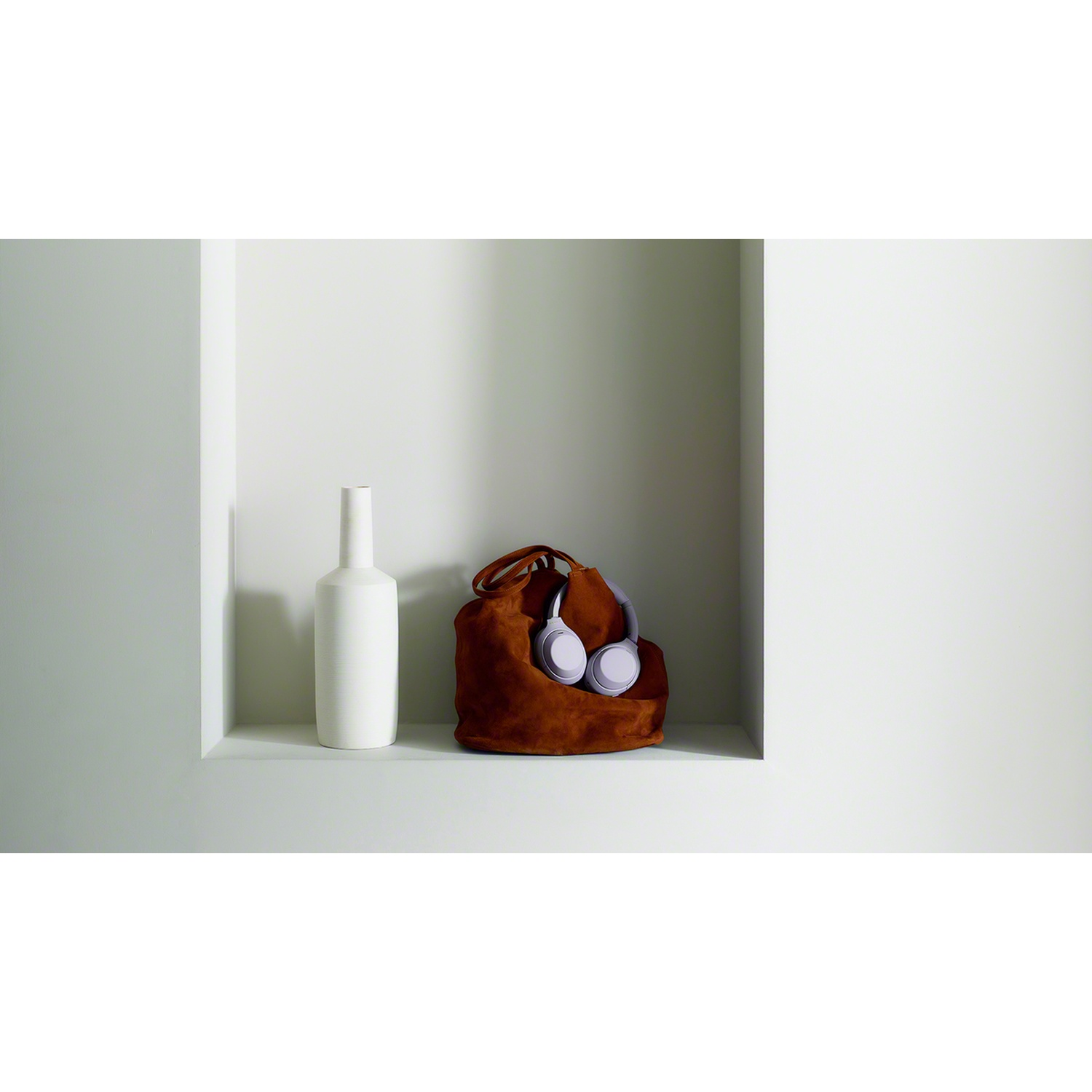Un vase blanc et un sac en daim marron avec des écouteurs violets dans une niche murale blanche.