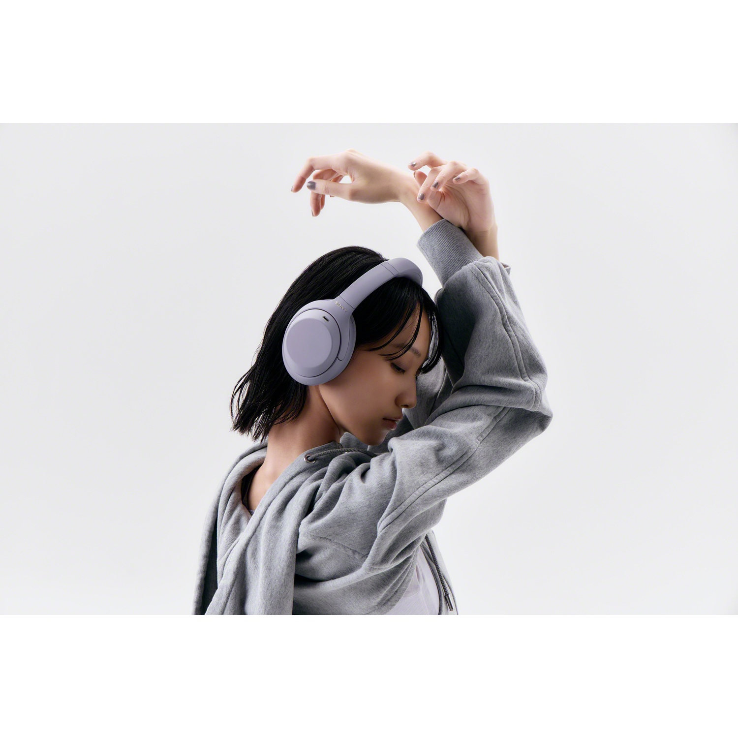 Une femme en sweat gris avec un casque violet lève les bras au-dessus de sa tête sur fond clair.