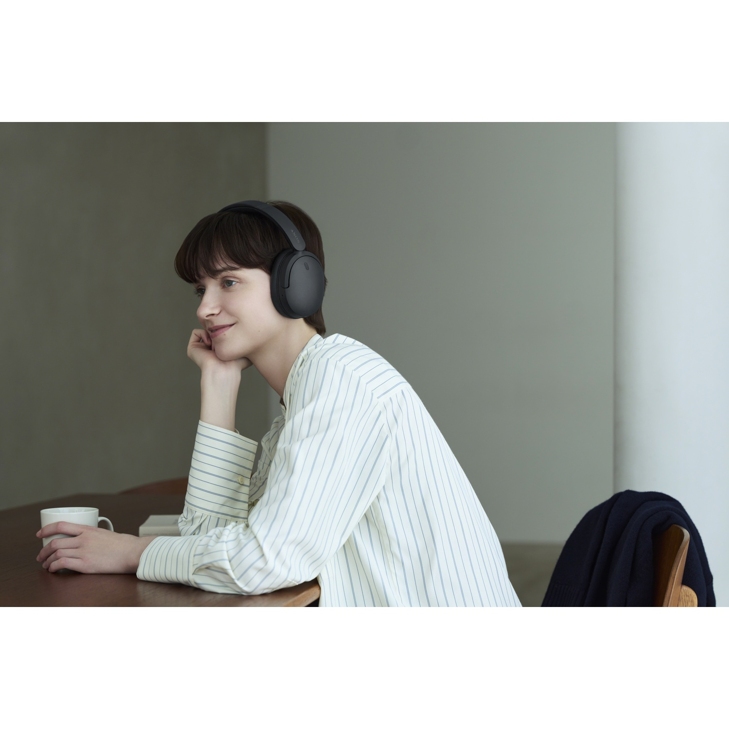 Une femme portant un casque noir est assise à une table en bois et tient une tasse blanche.