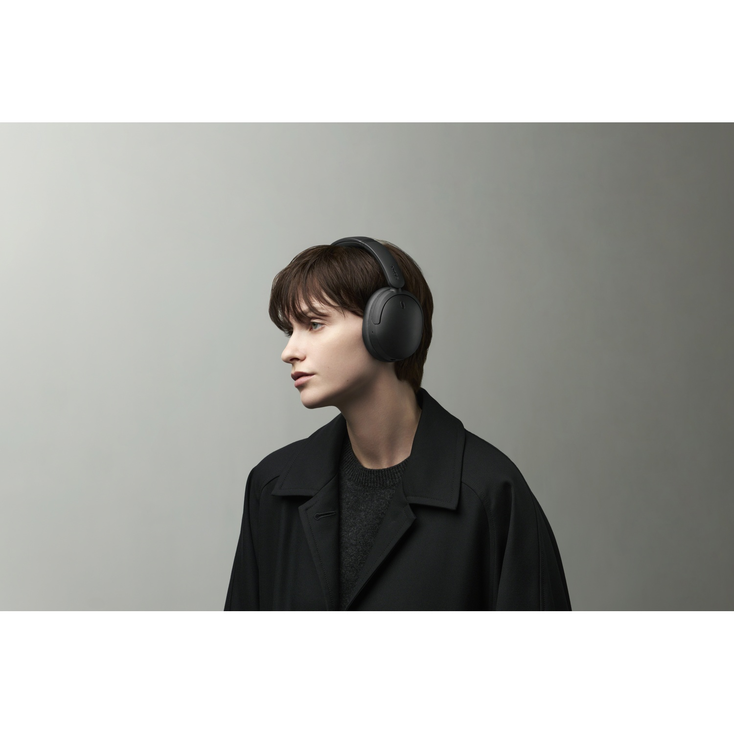 Une personne aux cheveux bruns courts porte un casque noir et un manteau sombre sur fond gris.