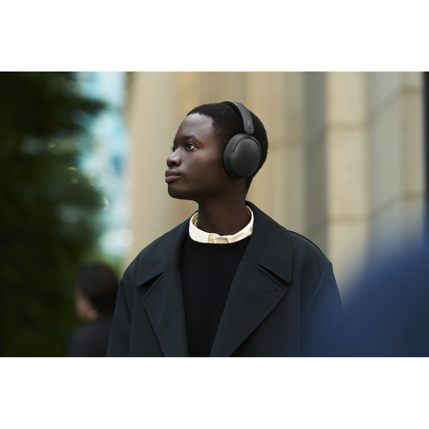Un jeune homme portant un casque sombre, un manteau noir et une chemise blanche regarde sur le côté à l'extérieur.
