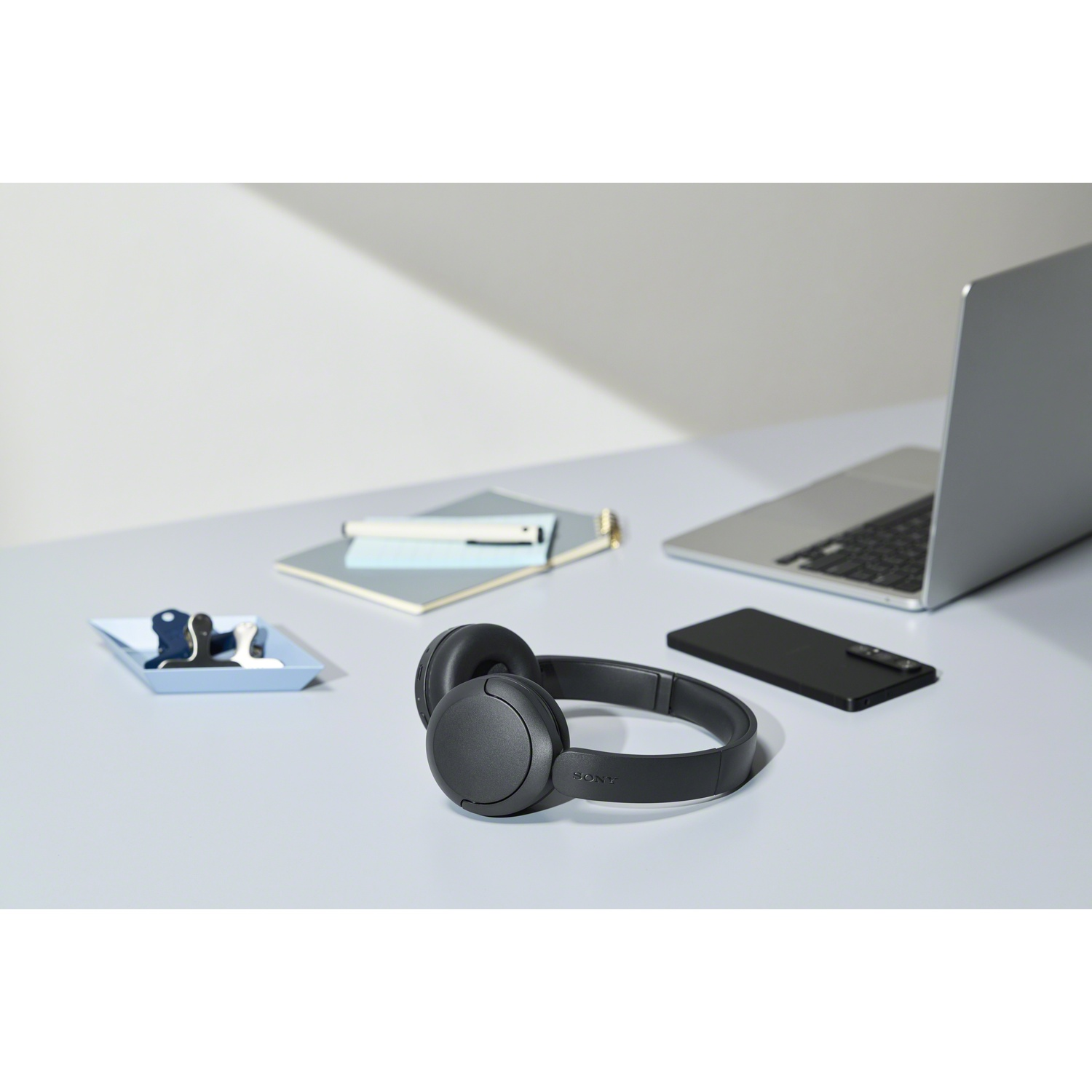 Casque sans fil Sony noir sur un bureau bleu clair avec un ordinateur portable, un smartphone et un carnet.