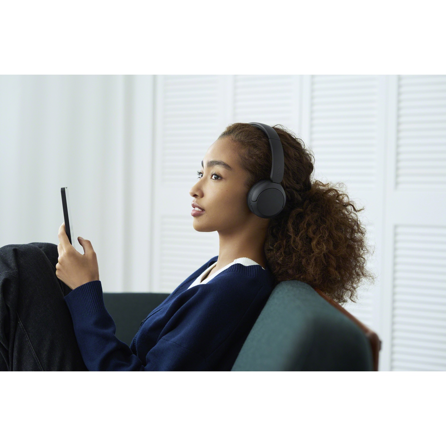 Une jeune femme aux cheveux bouclés et casquée est assise sur un canapé, tenant une tablette.