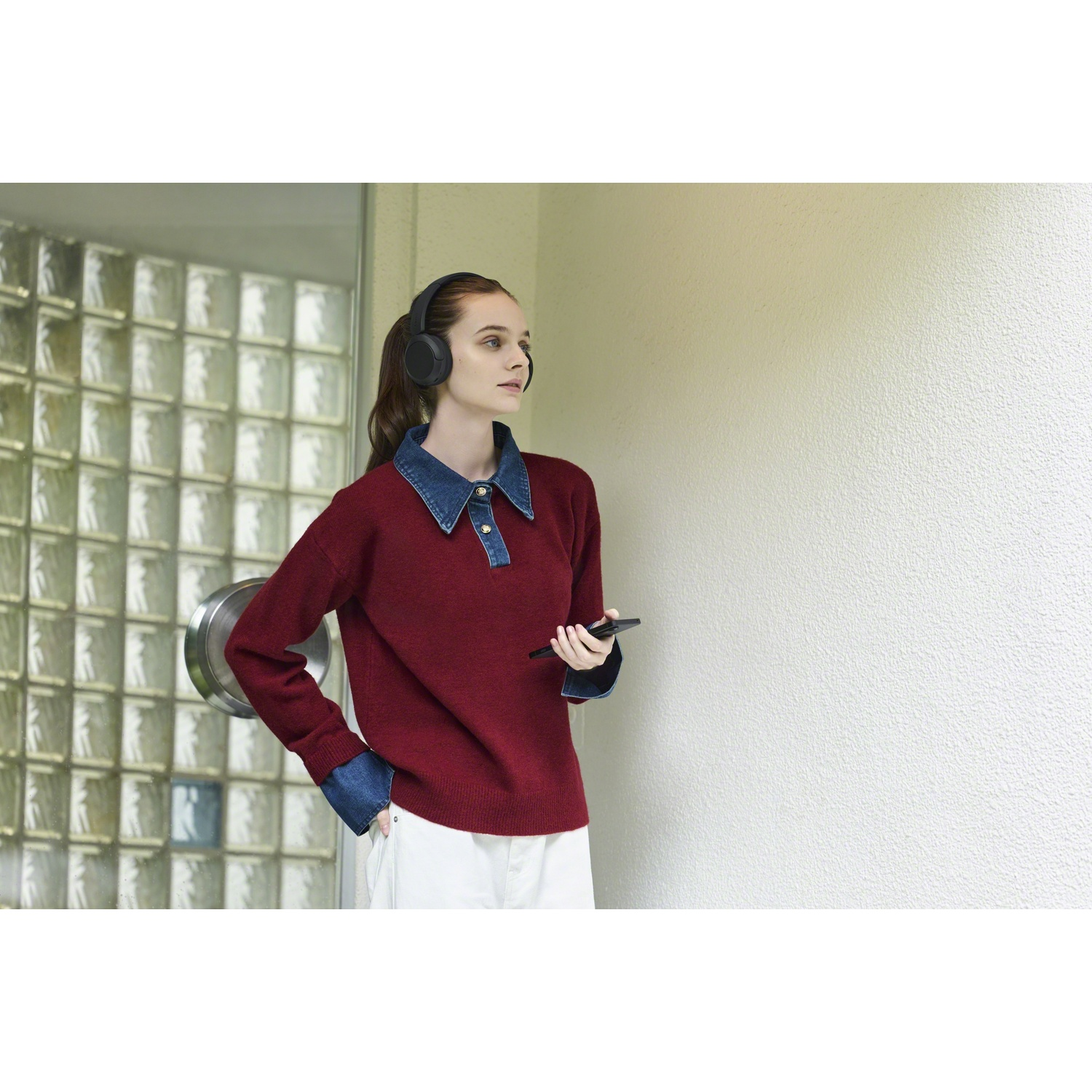 Une jeune femme en pull rouge, portant un casque et tenant un smartphone, se tient à l'extérieur.
