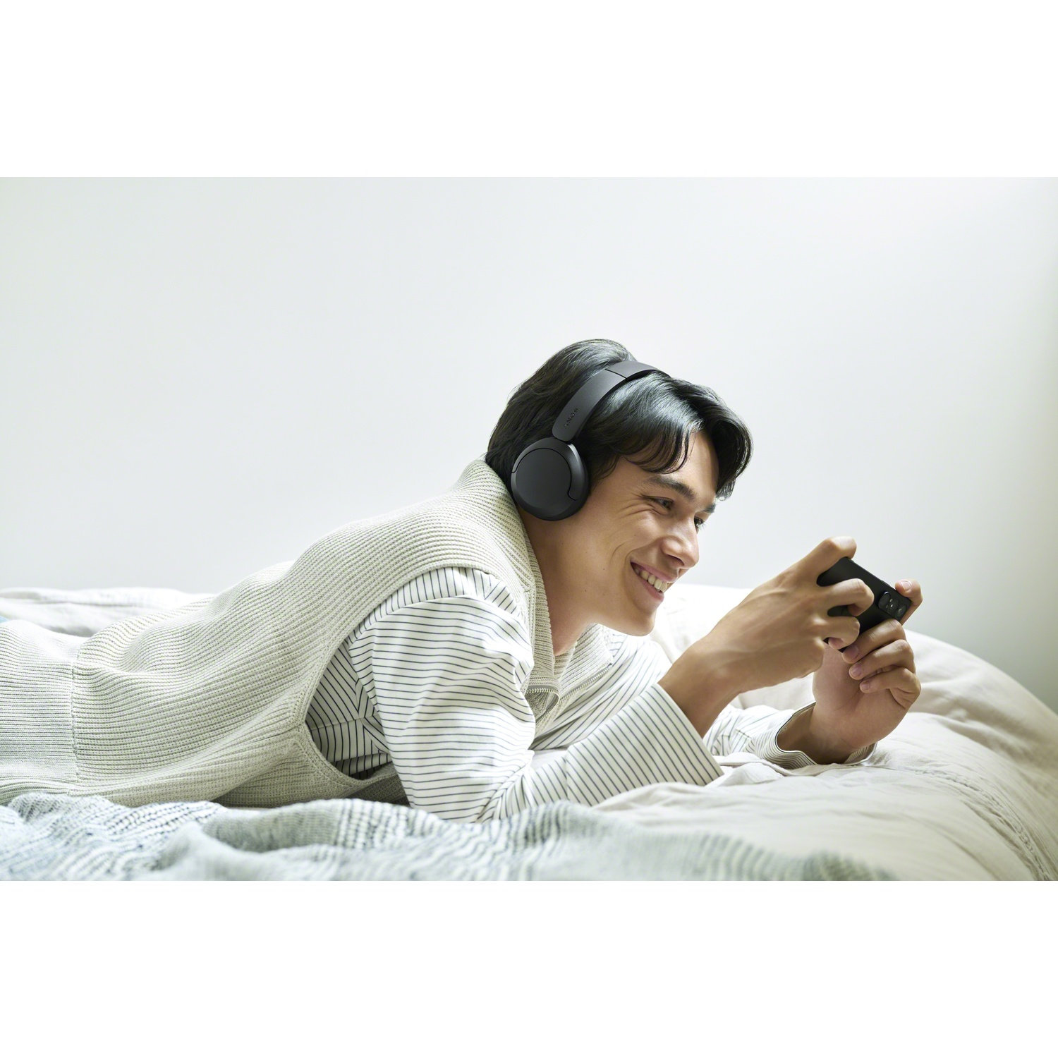 Un jeune homme portant un casque est étendu sur un lit, souriant en jouant sur son smartphone.