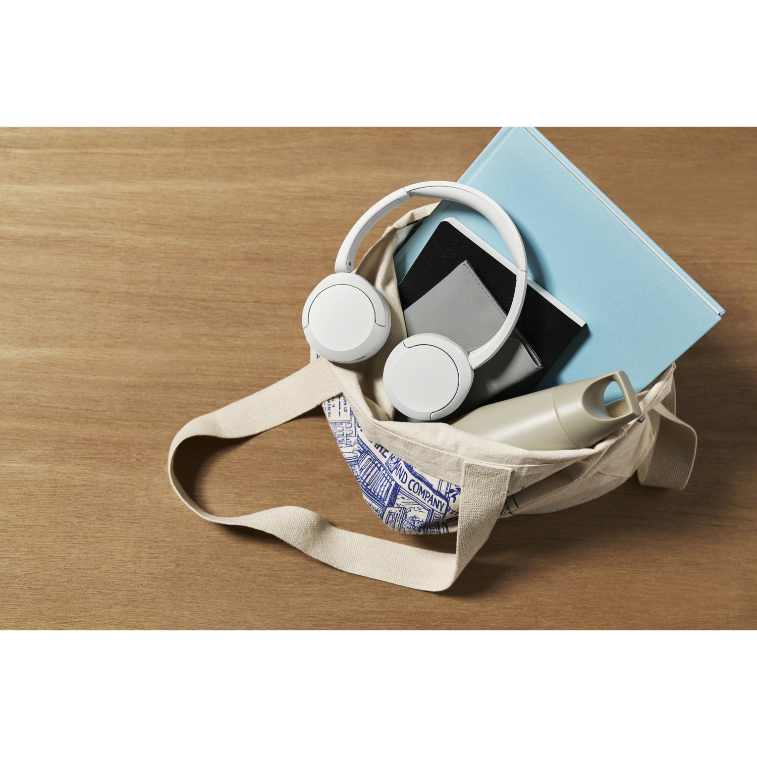 Un sac en tissu sur un sol en bois contient des écouteurs blancs, un livre bleu, des carnets et une bouteille.