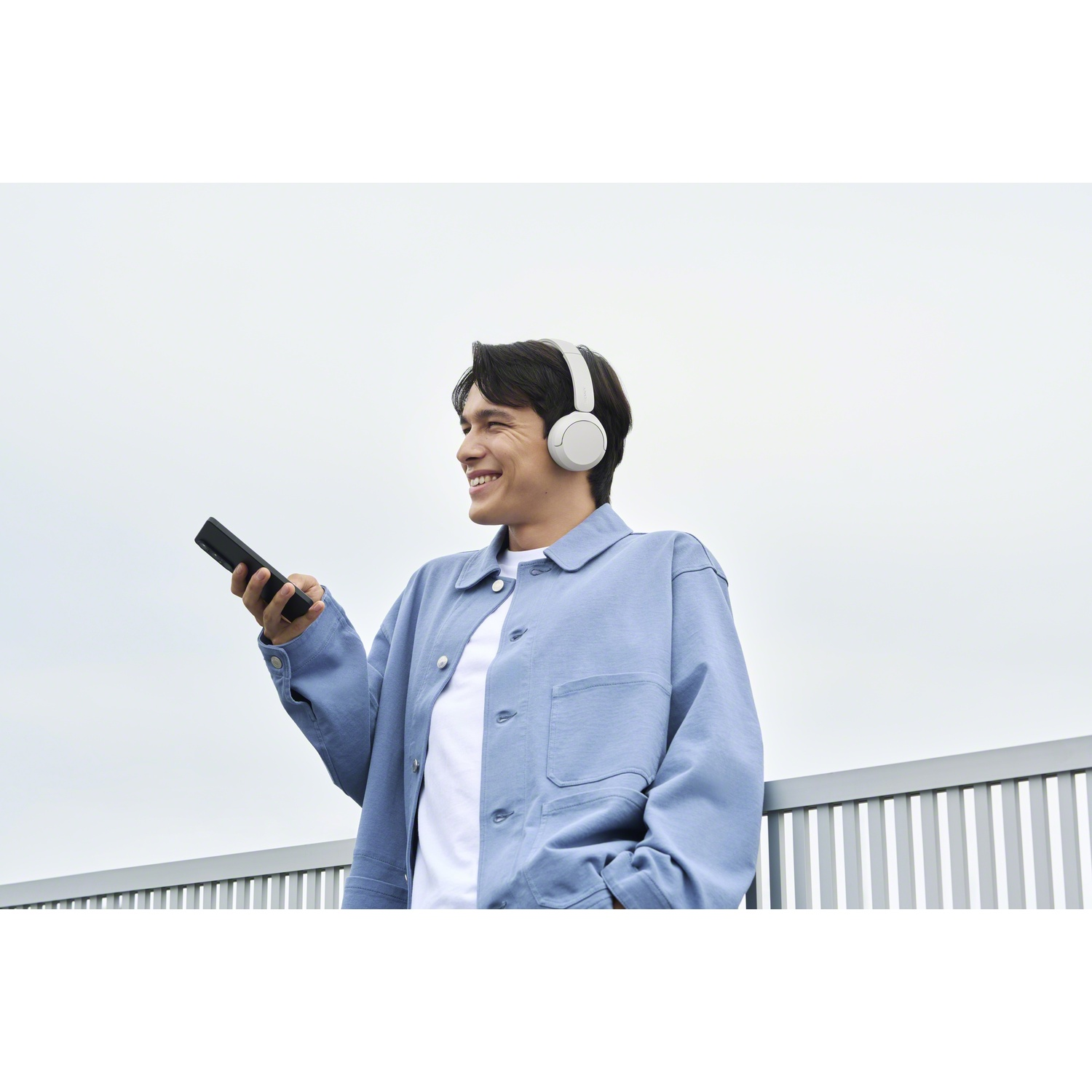 Un homme souriant en veste bleue et casque blanc tient un smartphone près d'un balcon.