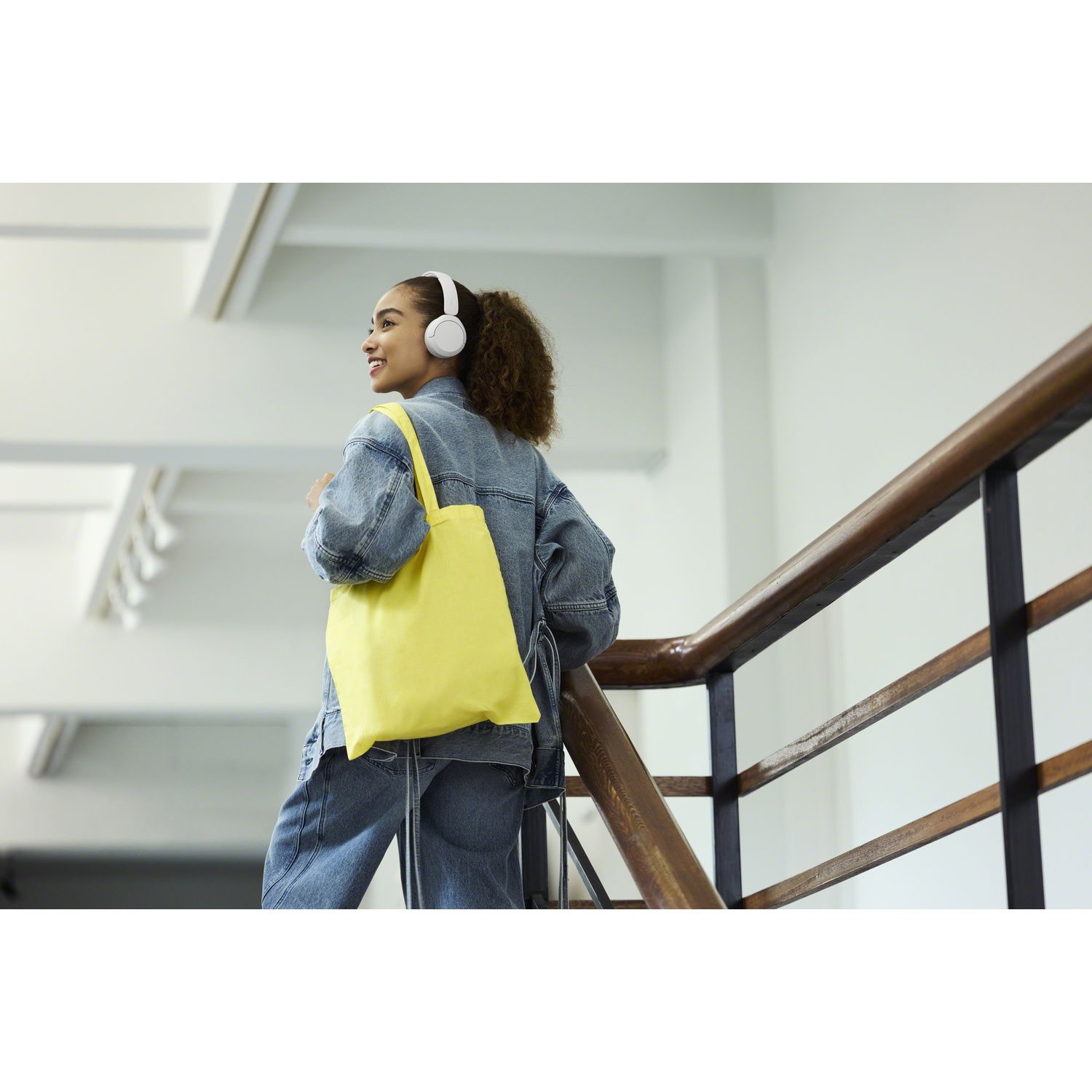 Une femme souriante en jean avec un casque blanc et un sac jaune monte des escaliers en bois.