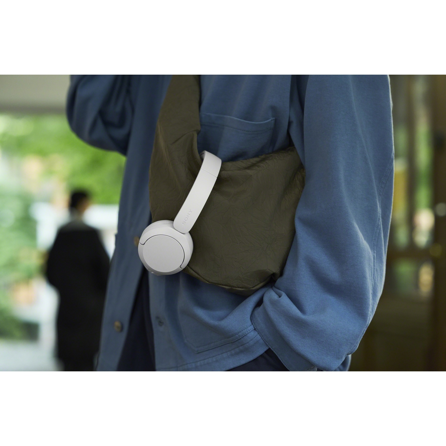 Une personne en chemise bleue porte un sac bandoulière vert avec des écouteurs blancs.