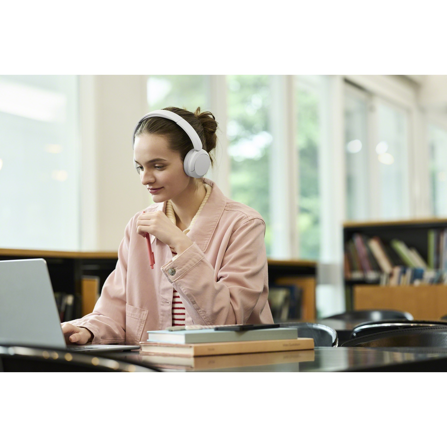 Une jeune femme avec un casque blanc est assise à un bureau avec un ordinateur portable, tenant un stylo.