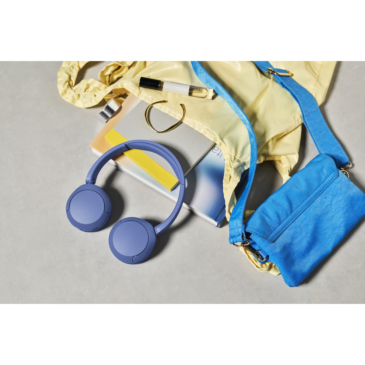 Casque bleu, sac jaune, livre, bracelet doré et rouge à lèvres sur fond gris.