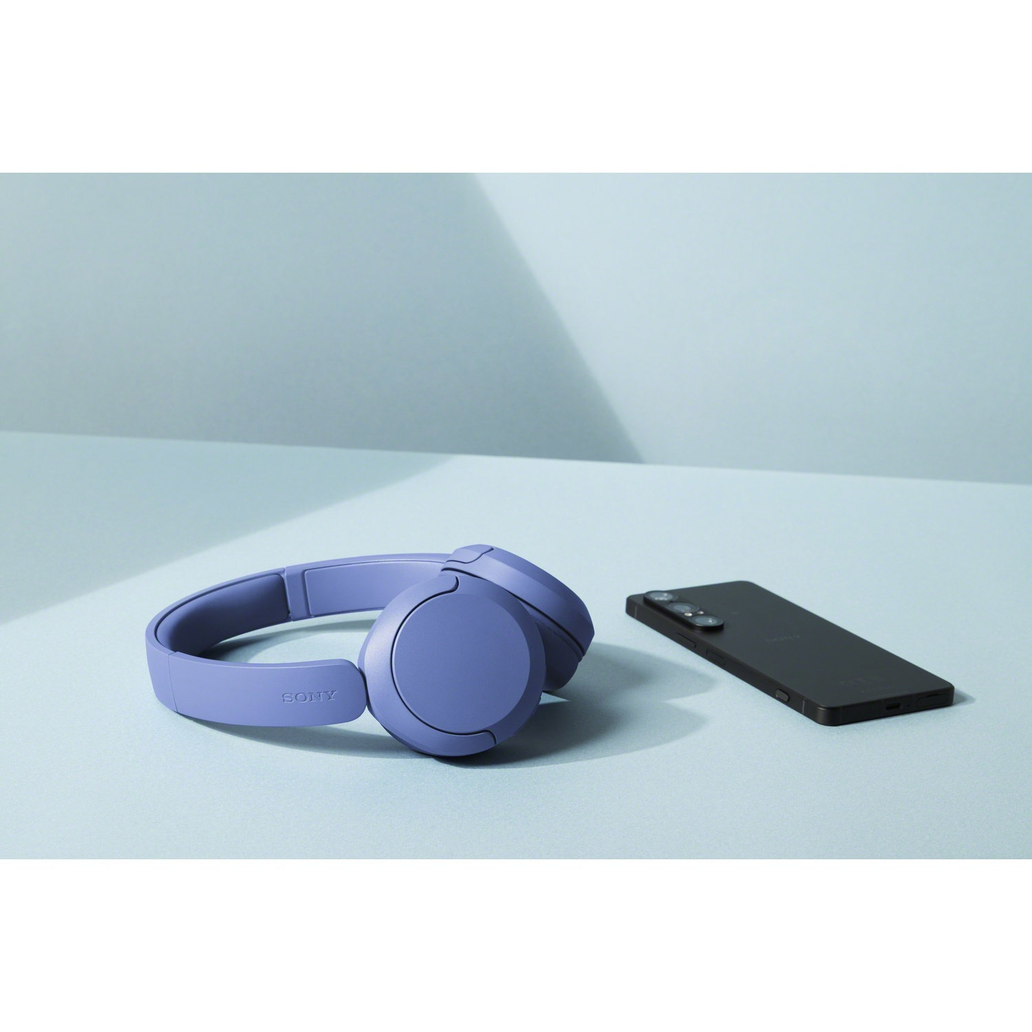 Un casque Sony violet et un smartphone noir reposent sur une table bleu clair devant un mur bleu clair.