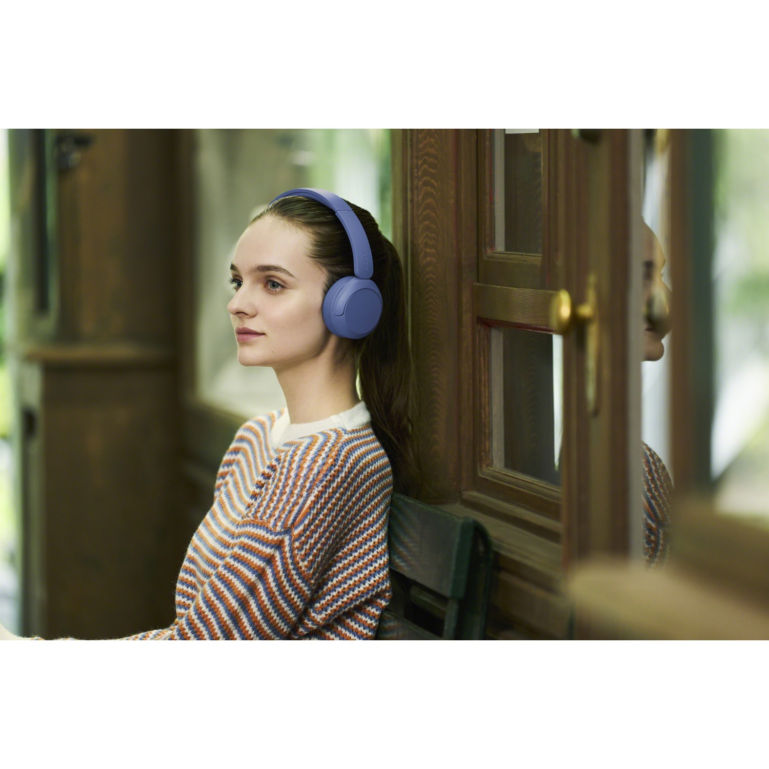 Une jeune femme en pull rayé et casque bleu est assise près d'une fenêtre à cadre en bois.